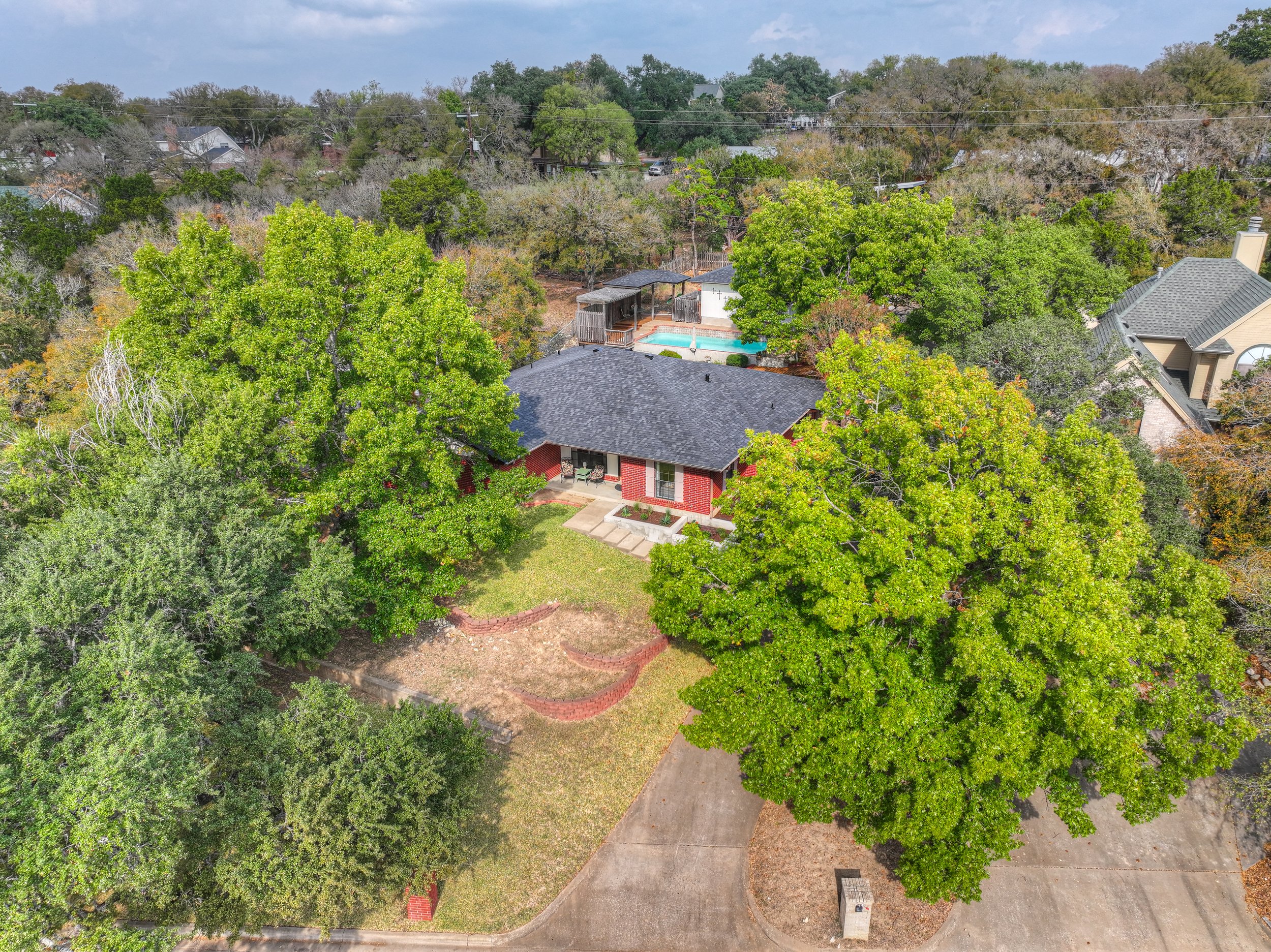 Aerial view of a house with a gray roof, surrounded by lush green trees, a concrete driveway, and a yard with a small garden bed. In the backyard, there's a swimming pool with a wooden deck and a shaded area.