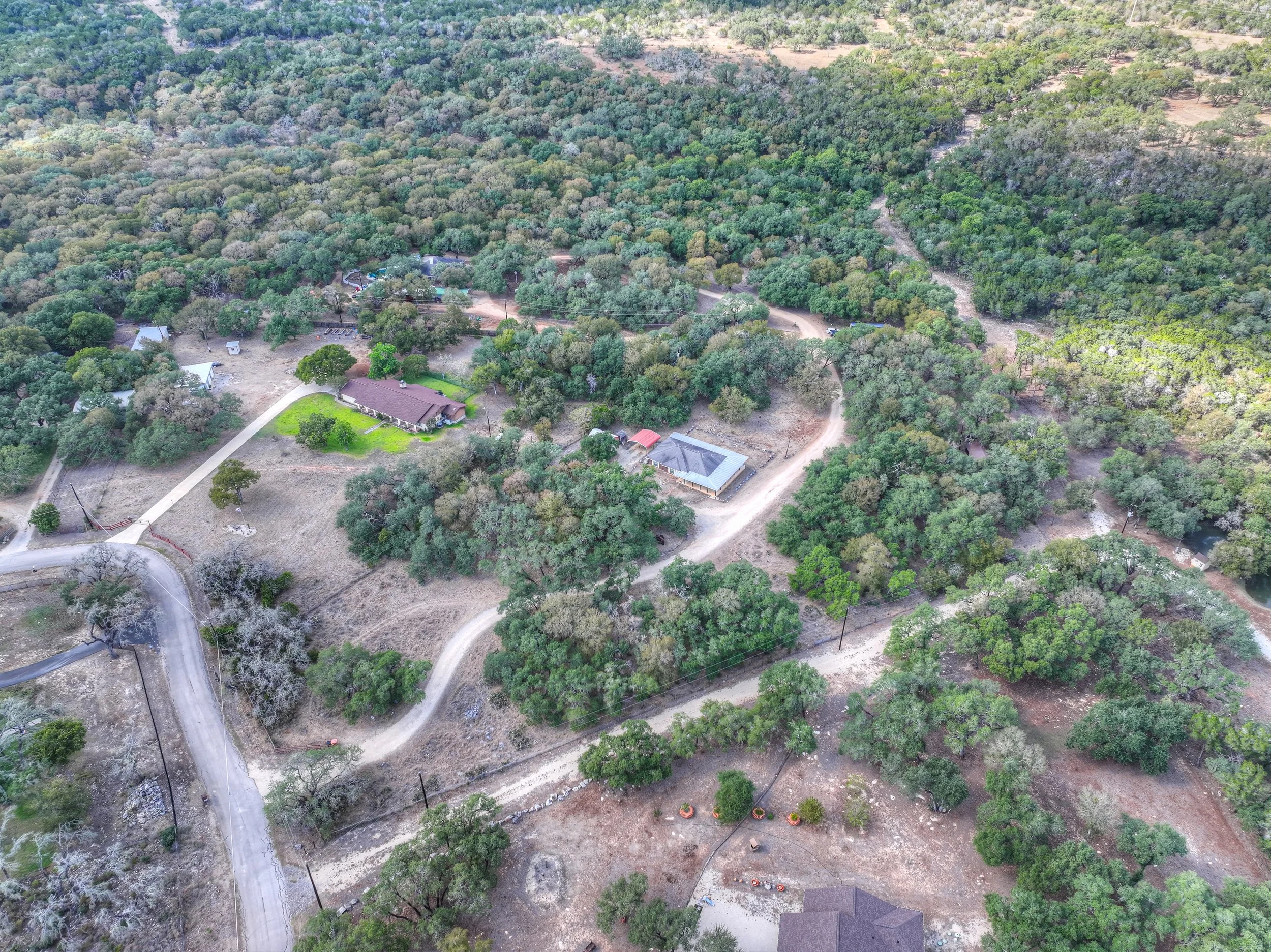 An aerial view of a rural area with trees, dirt roads, and scattered houses, including a large house with a gray roof and a porch, surrounded by greenery.