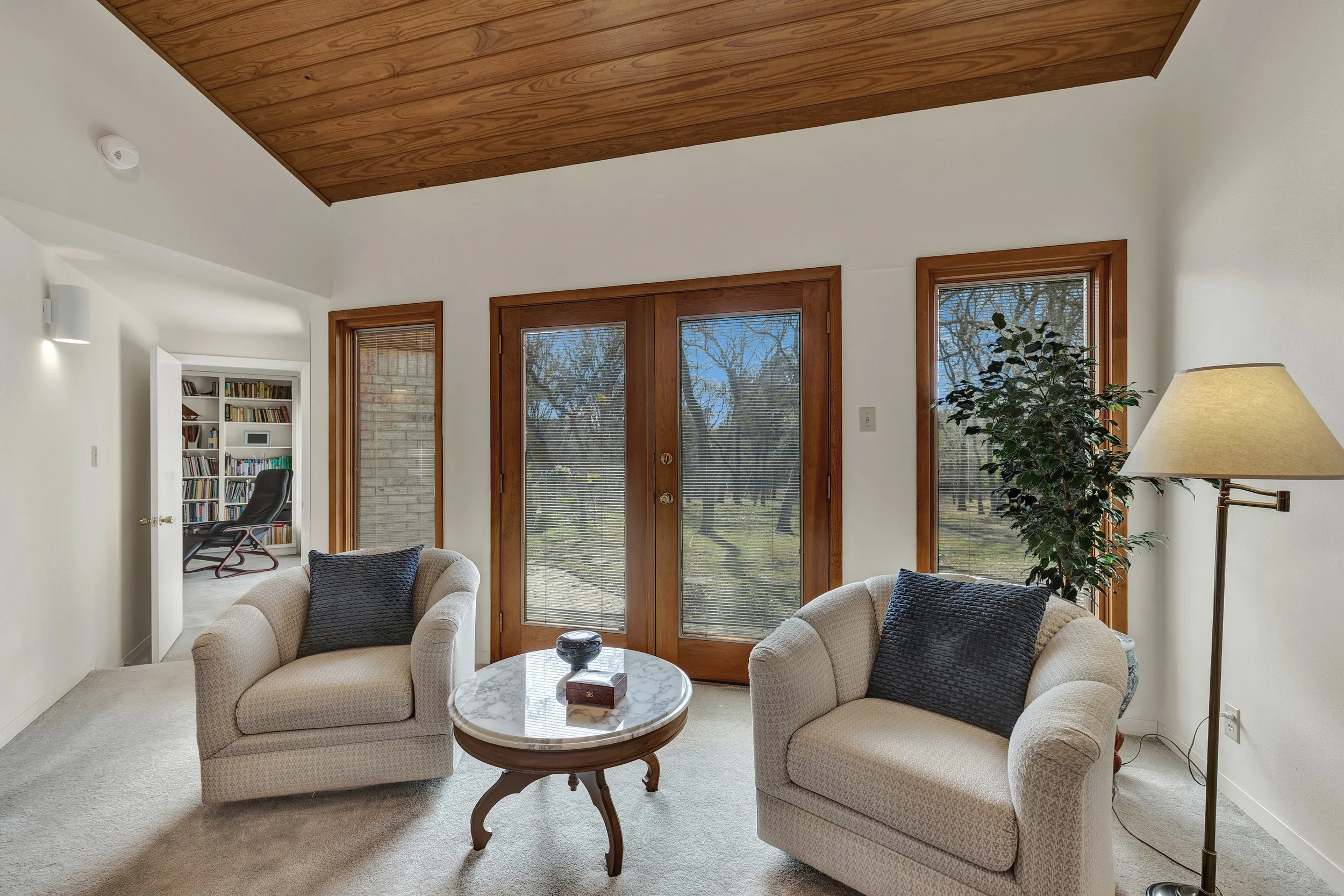 Living room with two beige armchairs with black pillows, a small marble-top coffee table, a wood-paneled ceiling, glass doors, a tall floor lamp, and a large potted plant. In the background, there is a doorway to a study with a bookshelf and a black 