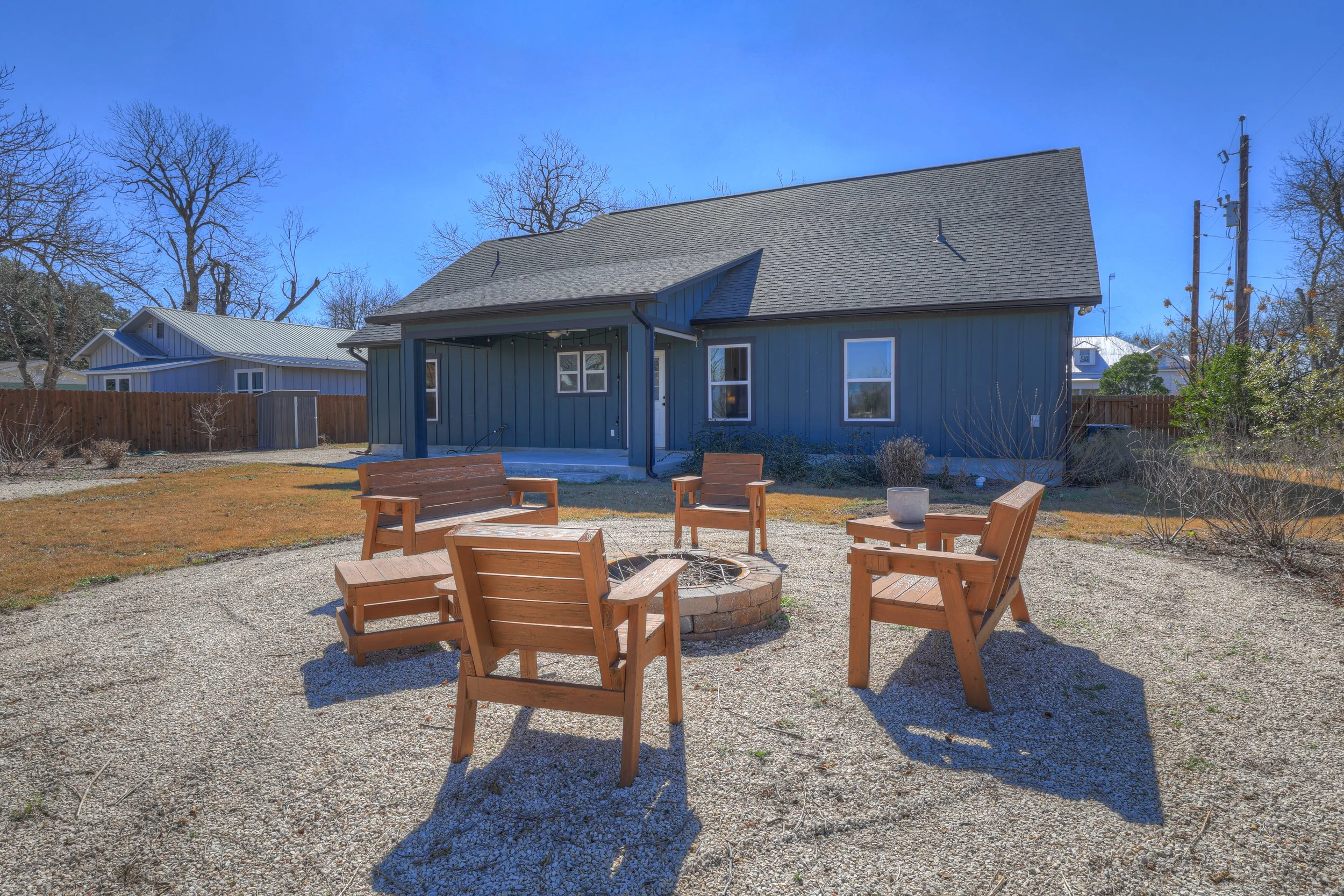 Backyard with a fire pit surrounded by five wooden chairs, a grassy area, and a blue house with a porch and three windows.