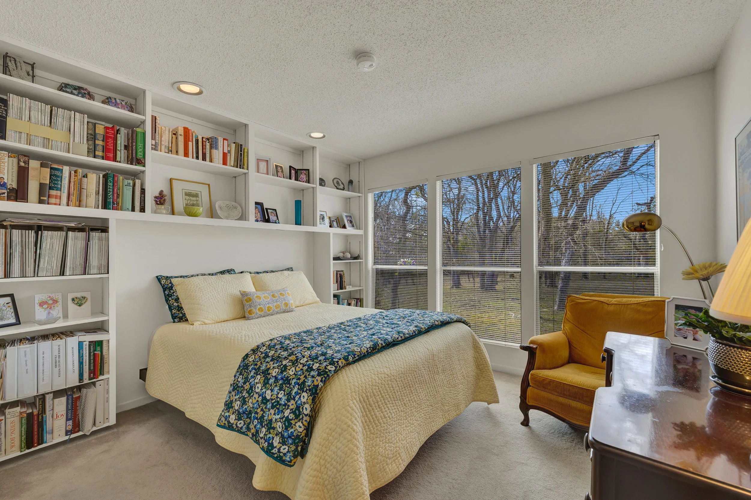 A bedroom with a bed covered in a yellow quilt, floral blanket, and pillows, next to a white bookshelf filled with books and framed photos, near large windows showing leafless trees outside, with a yellow armchair and a wooden dresser with a table la