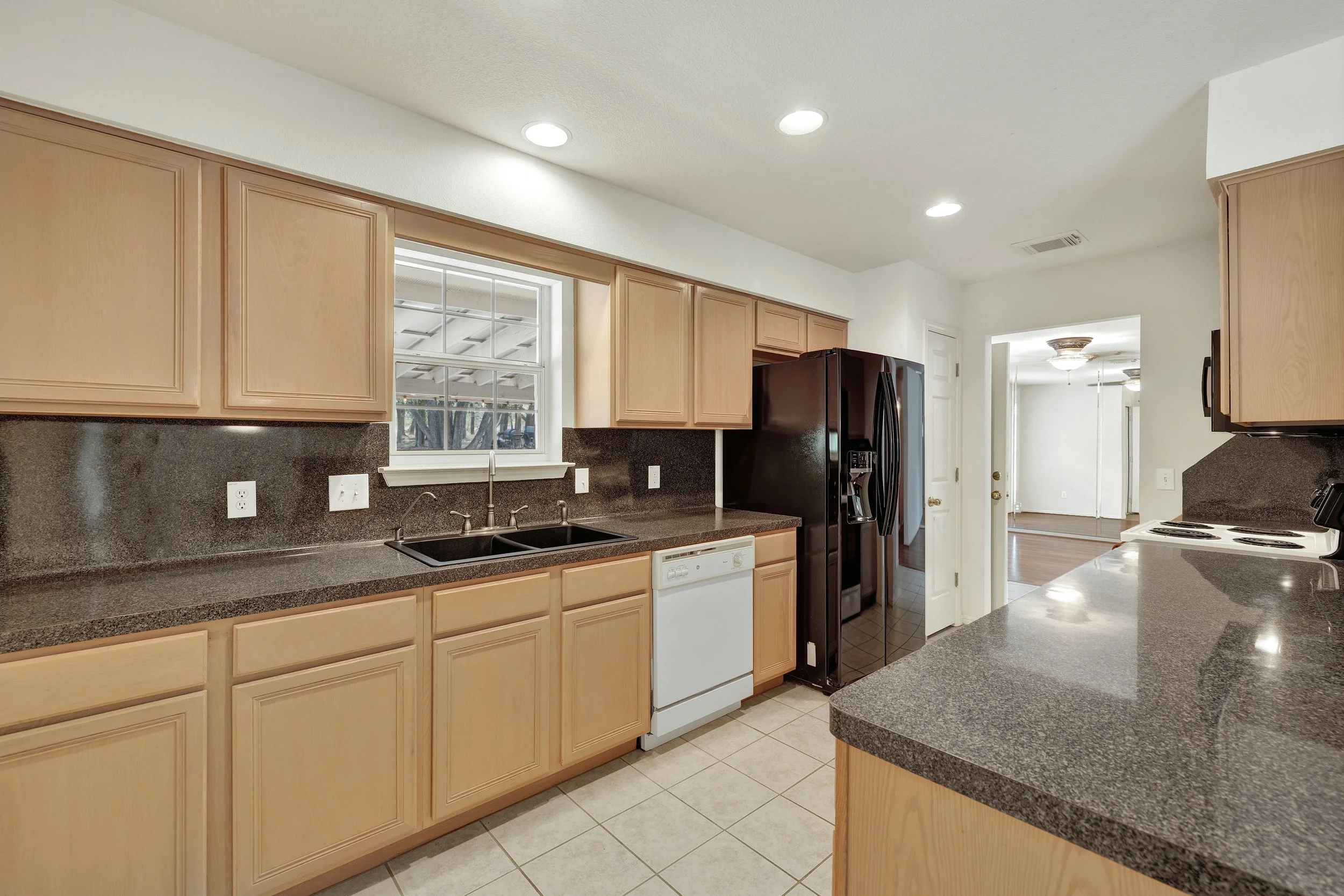Kitchen with light wood cabinets, black refrigerator, white dishwasher, black countertops, and a window over the sink.