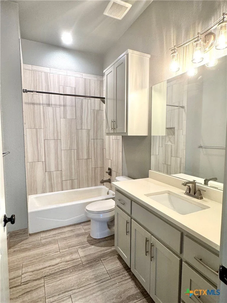 A modern bathroom with a bathtub and shower, toilet, white vanity with a sink, mirror, and cabinet, and wood-look floor and wall tiles.