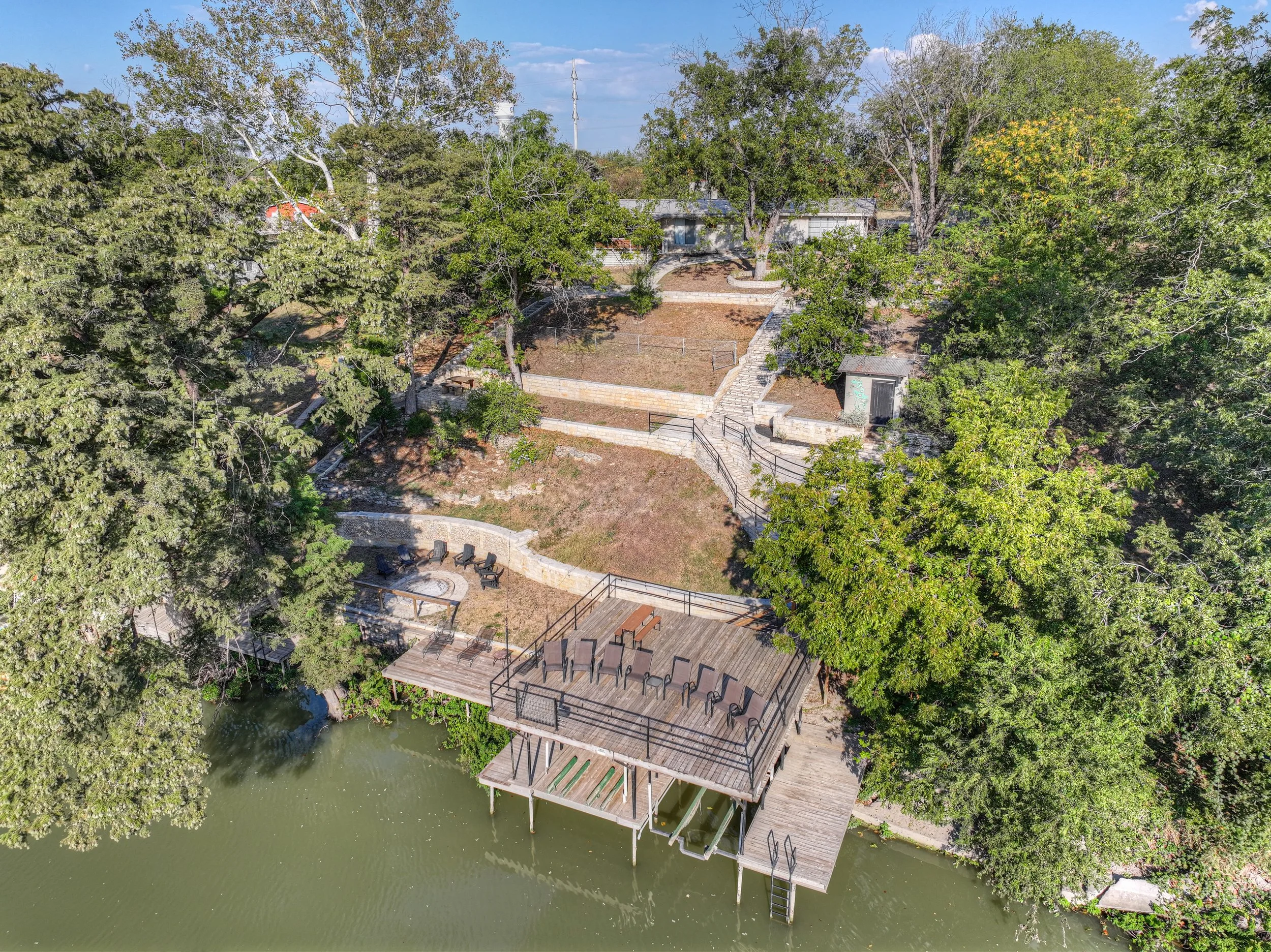Aerial view of a lakeside backyard with a deck and outdoor seating, surrounded by trees, with stairs leading up to a terraced yard with more seating and pathways.