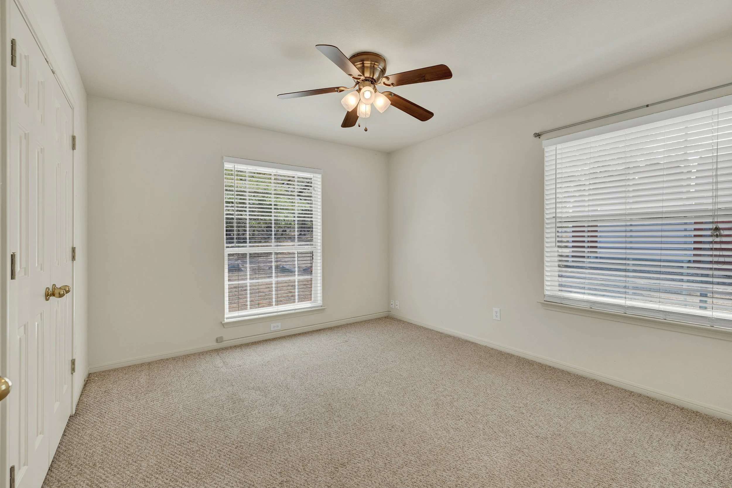 Empty bedroom with beige carpet, white walls, two windows with blinds, ceiling fan with wooden blades and light fixtures.