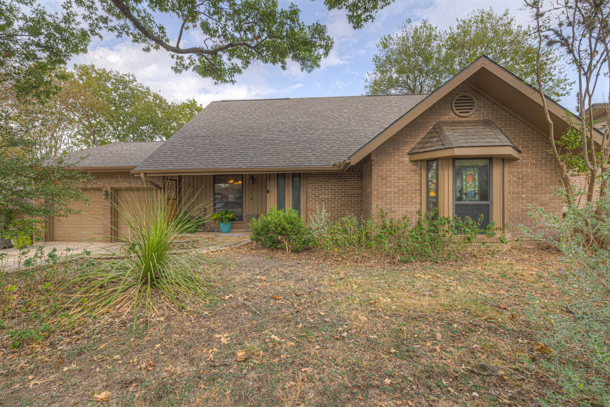 A single-story brick house with a gray shingled roof, a small front porch, and a bay window. There are trees and shrubs in the front yard, some dry grass and fallen leaves on the ground, and a blue pot with a green plant near the front door.