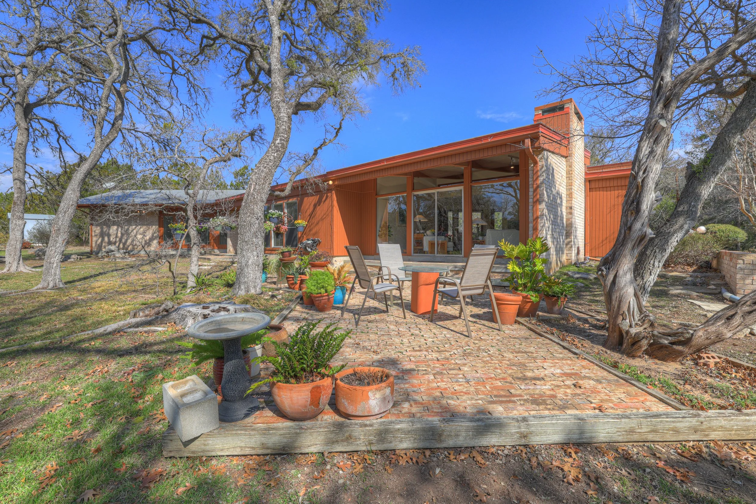 Backyard patio with brick flooring, potted plants, outdoor dining set with four chairs, leafless trees, and a house with orange siding and large glass doors