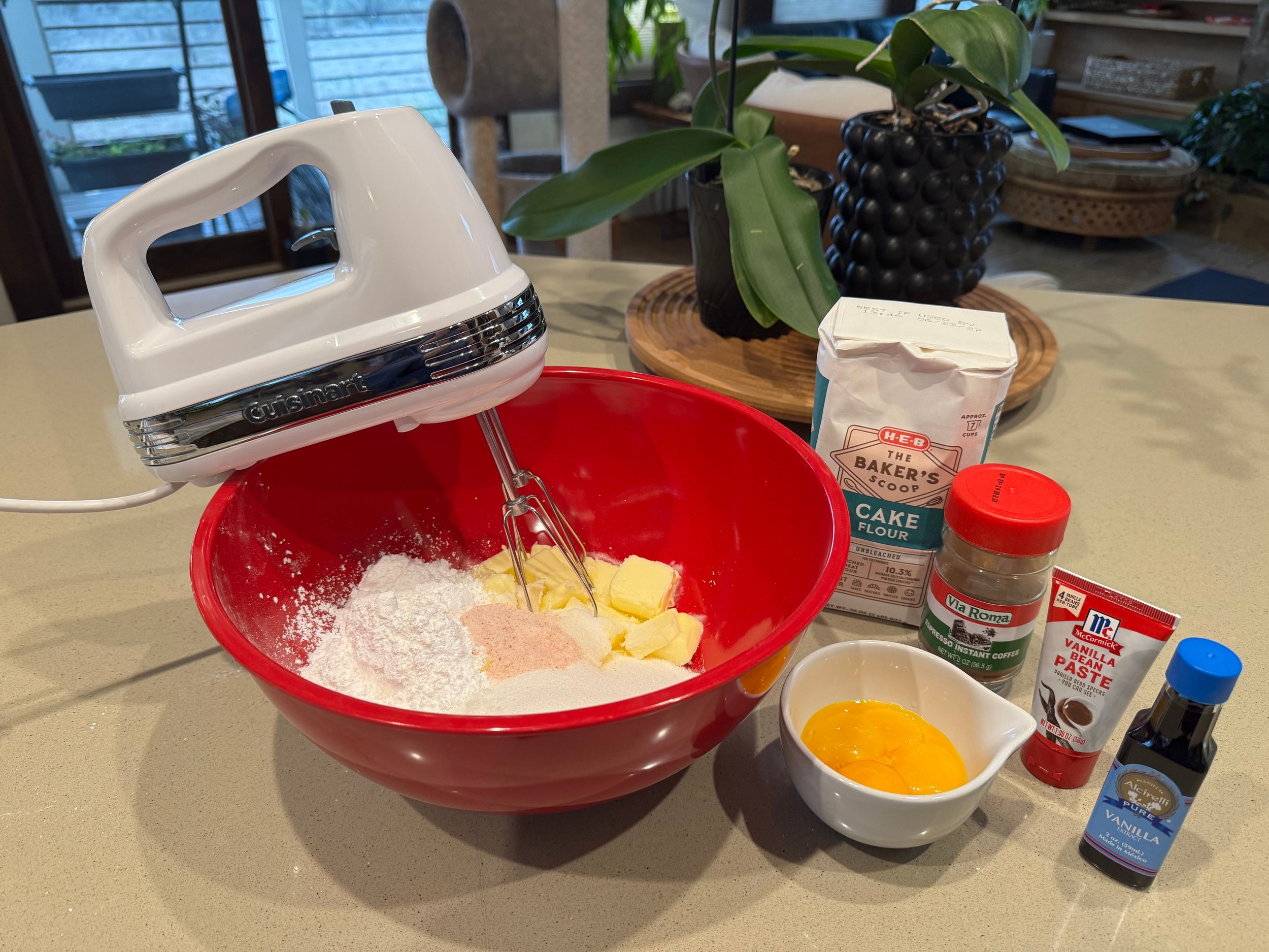 A red mixing bowl contains flour, butter, and sugar along with a hand mixer. Nearby are ingredients including a bowl with cracked eggs, a bag of cake flour, a jar of vanilla bean paste, a jar of instant coffee, and a bottle of vanilla extract on a kitchen counter. In the background, there are potted plants and a wooden tray.