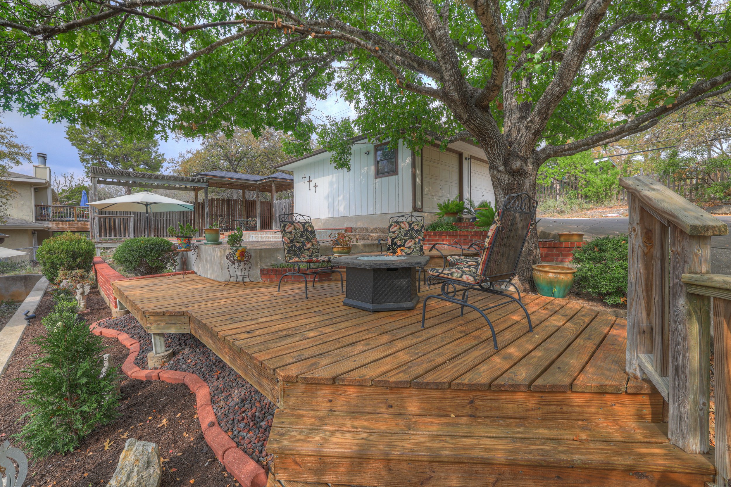 A backyard patio with a wooden deck, patio furniture, potted plants, and a large tree providing shade.
