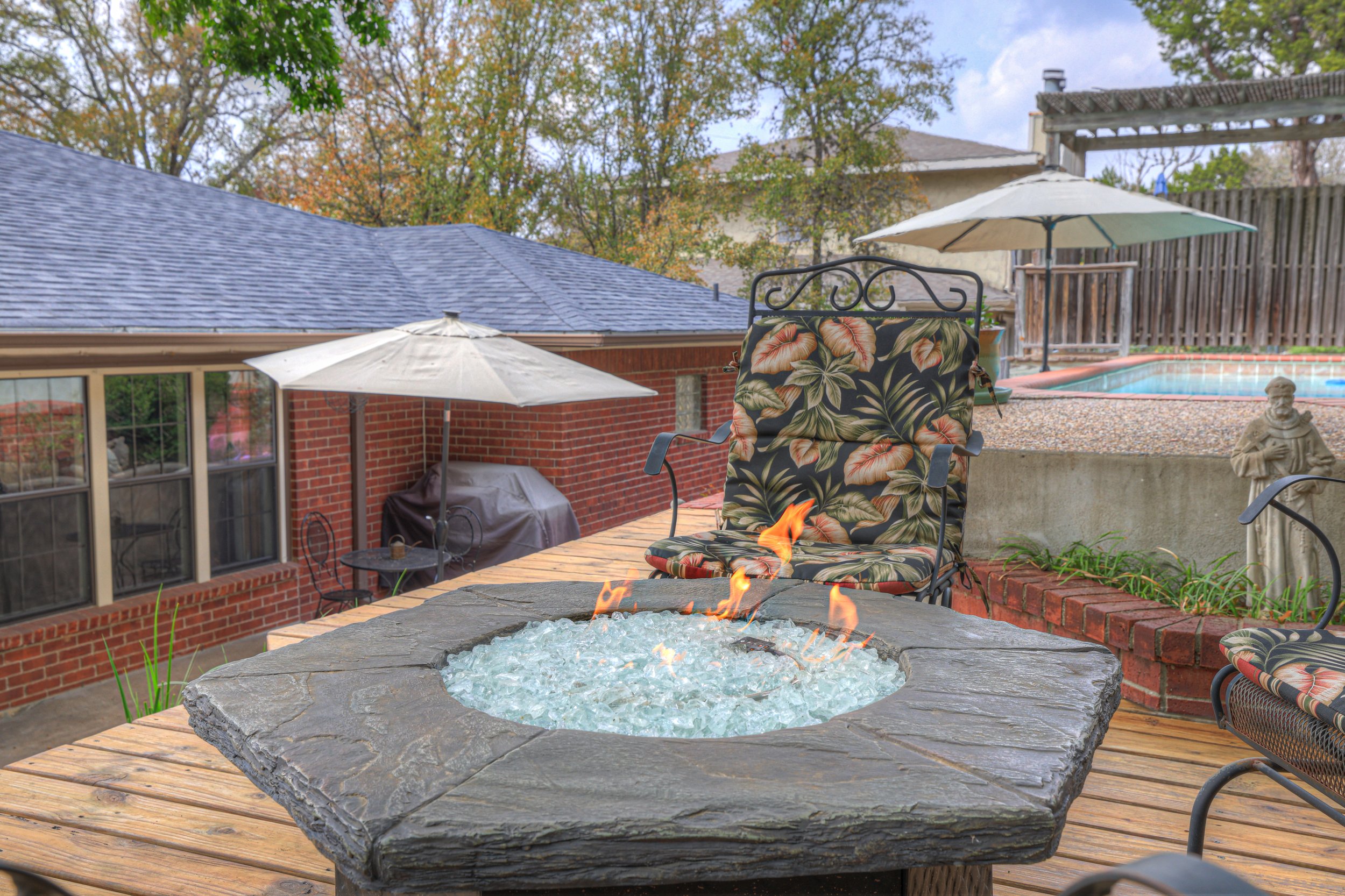 A backyard patio with a fire pit in the center, surrounded by outdoor chairs with floral cushions, with umbrellas providing shade, nearby a small pool, and trees in the background.
