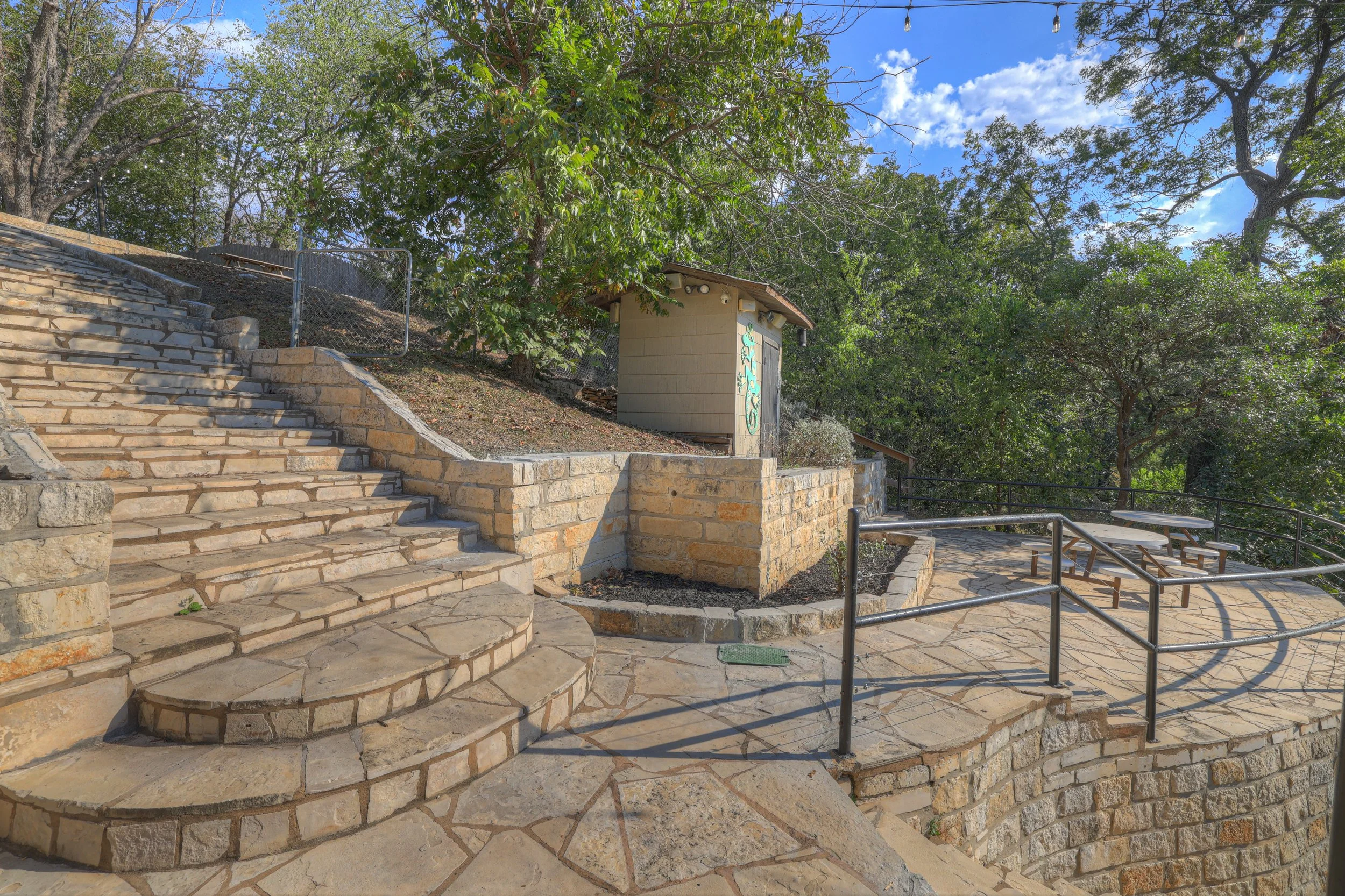 Stone steps and a small retaining wall with metal railings in an outdoor park setting, with trees and a small shed in the background.