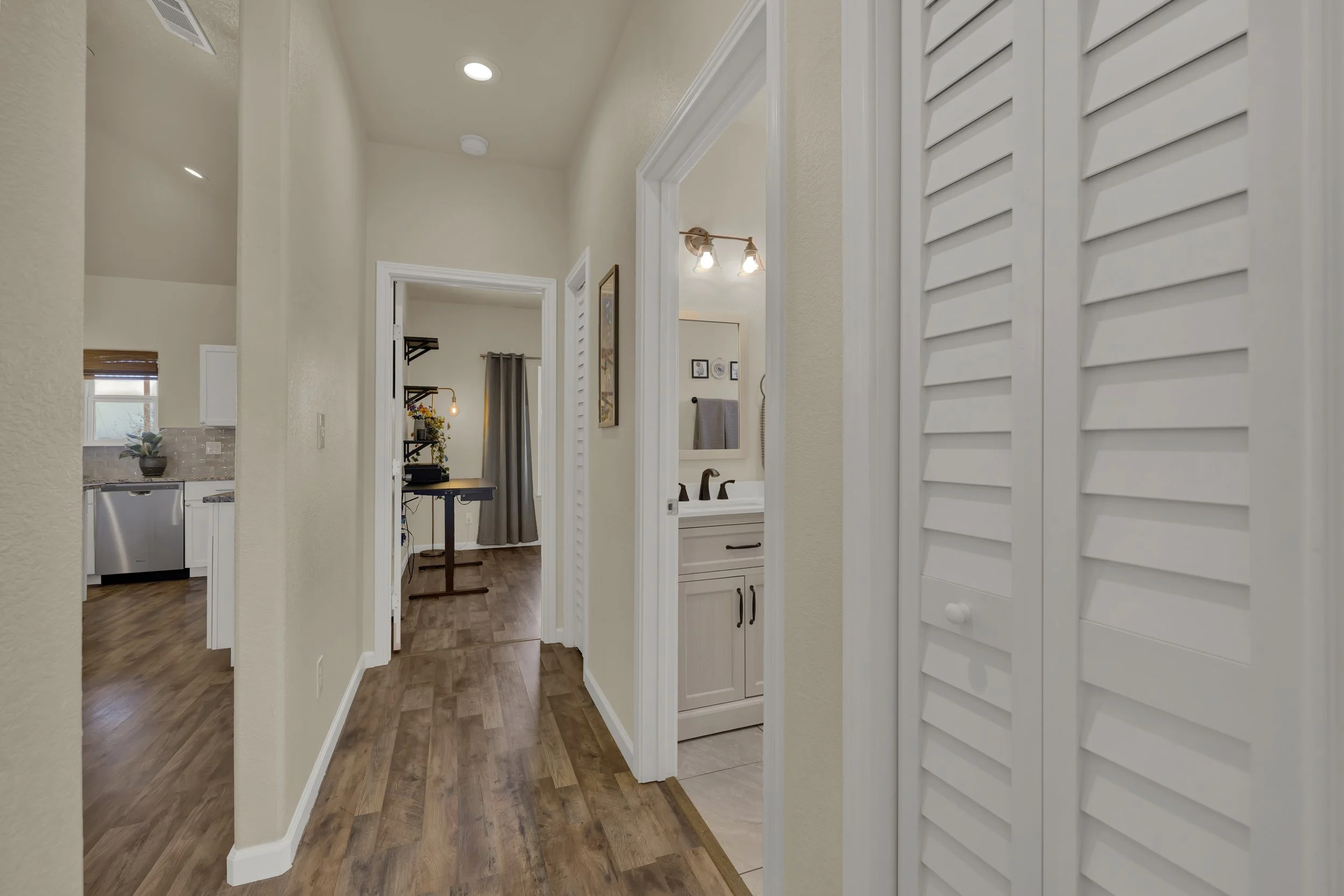 Interior view of a hallway in a home with hardwood floors, showing a bathroom with a vanity and mirror, a bedroom in the background with ceiling lights and curtains, and a kitchen area partially visible on the left.