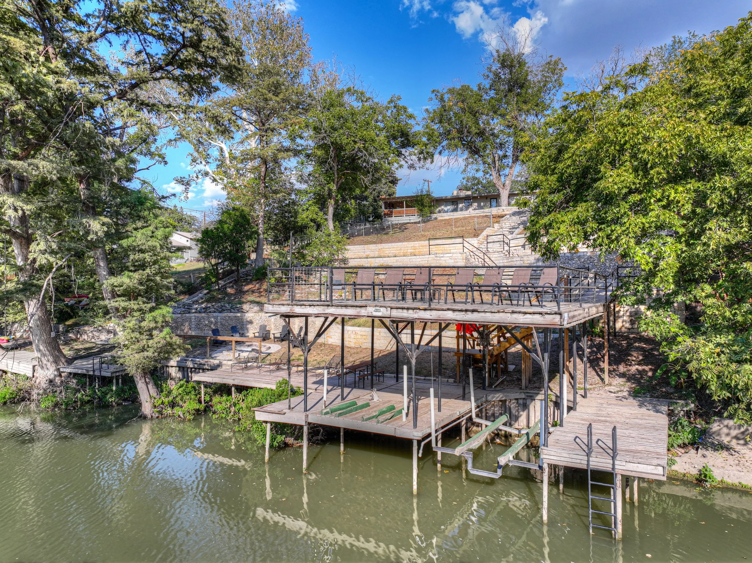 A scenic view of a riverfront area with multiple wooden decks and seating arrangements, surrounded by green trees and a blue sky with some clouds.