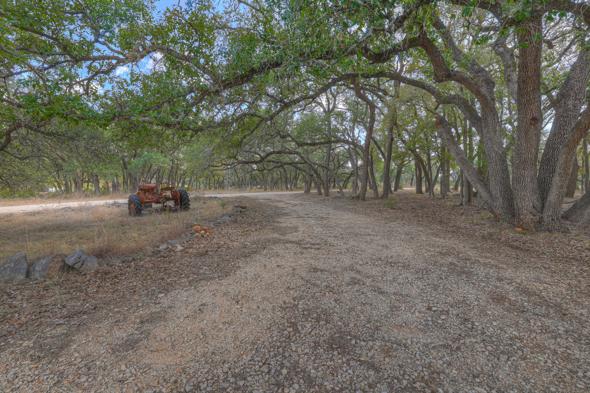 A dirt path through a wooded area with tall trees on both sides. An old rusty tractor is parked near the edge of the path on the left side.