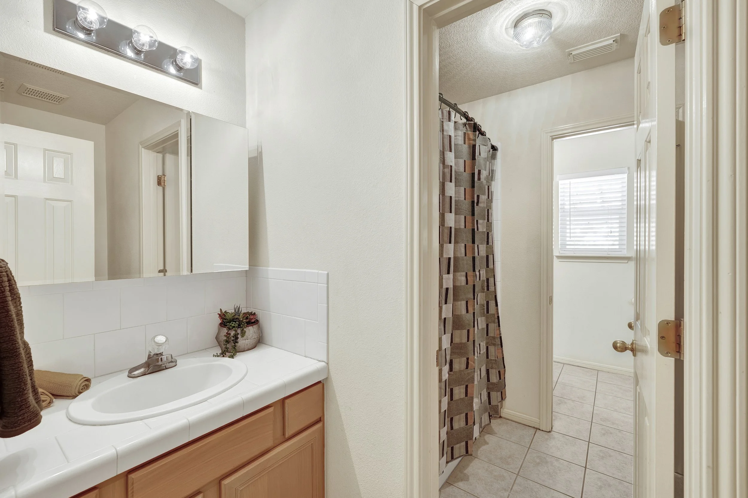 Bathroom with a white sink, mirror, and light fixture above, and a shower with a patterned curtain, a window with blinds, and tiled floor.
