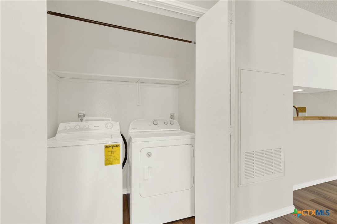 A laundry area with a white washing machine and dryer side by side against a white wall, with a shelf above and a narrow opening to the right.