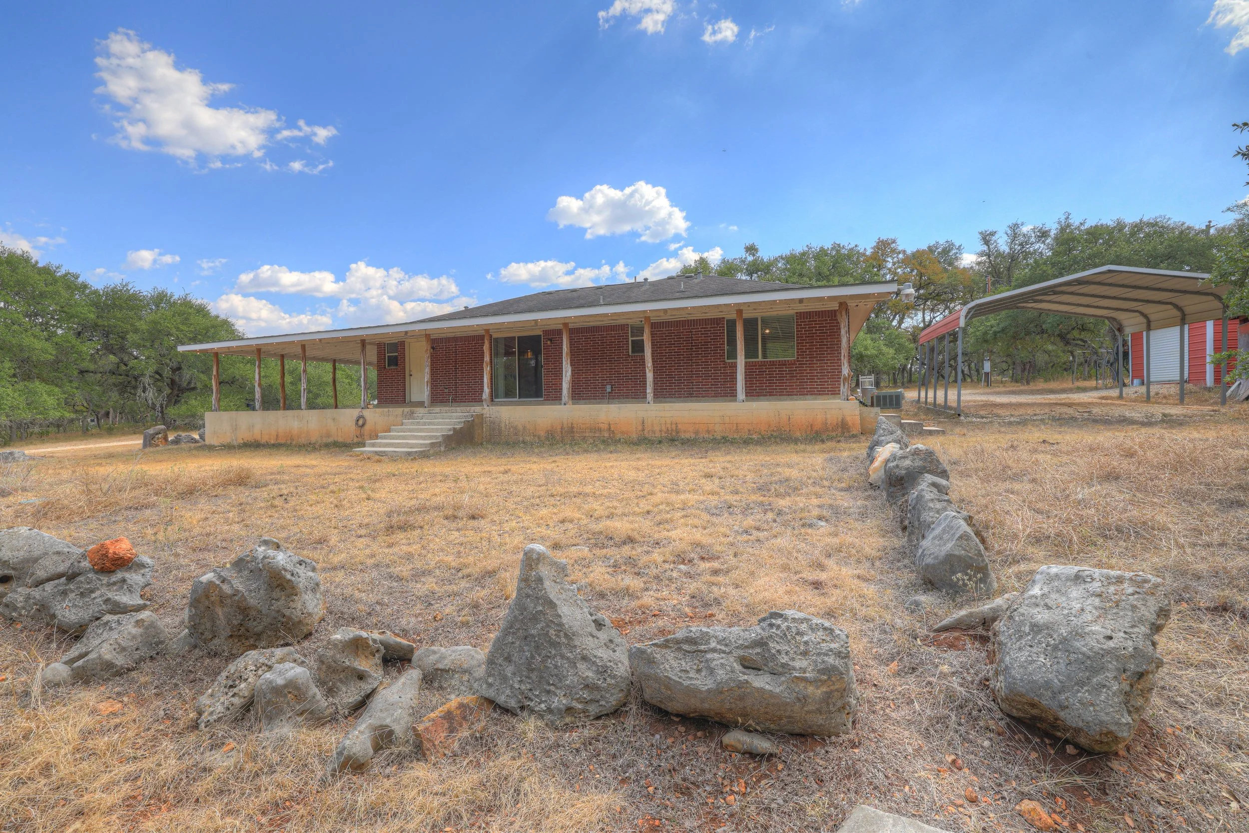 Front view of a single-story brick house with a large front porch and concrete steps leading up to it, set in a dry, grassy landscape with rocks along the edge. There is a carport to the right, a blue sky with scattered clouds, and some trees in the 