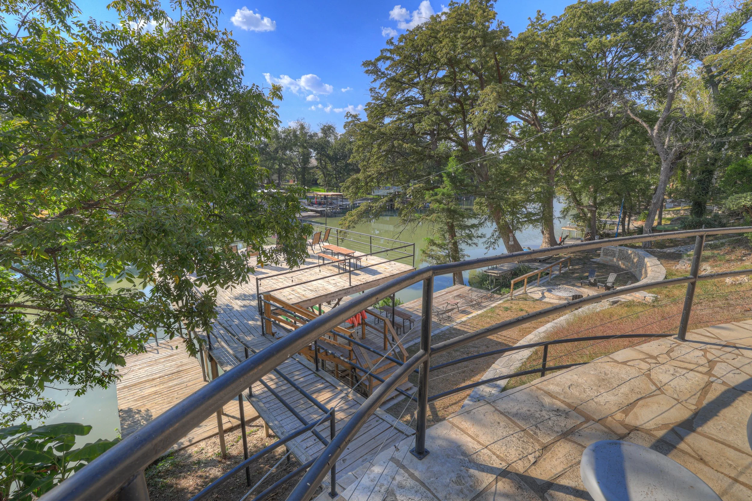 A view from a terrace overlooking a lake with trees and a wooden dock with chairs and benches on a sunny day
