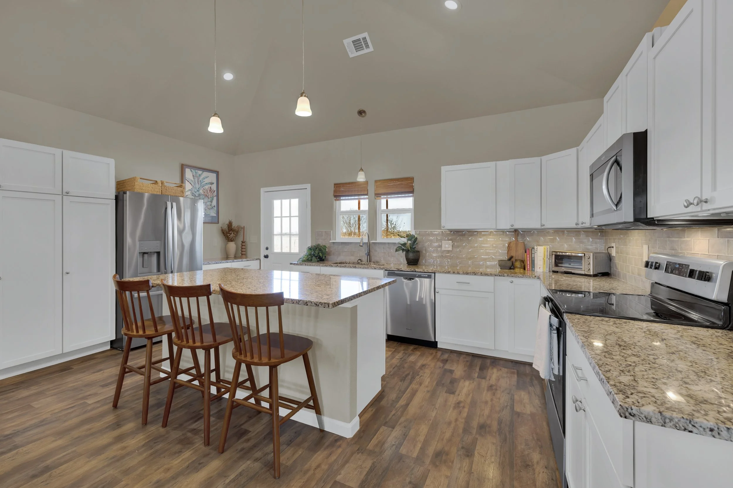 Modern kitchen with white cabinets, granite countertops, stainless steel appliances, wood flooring, and three wooden bar stools.