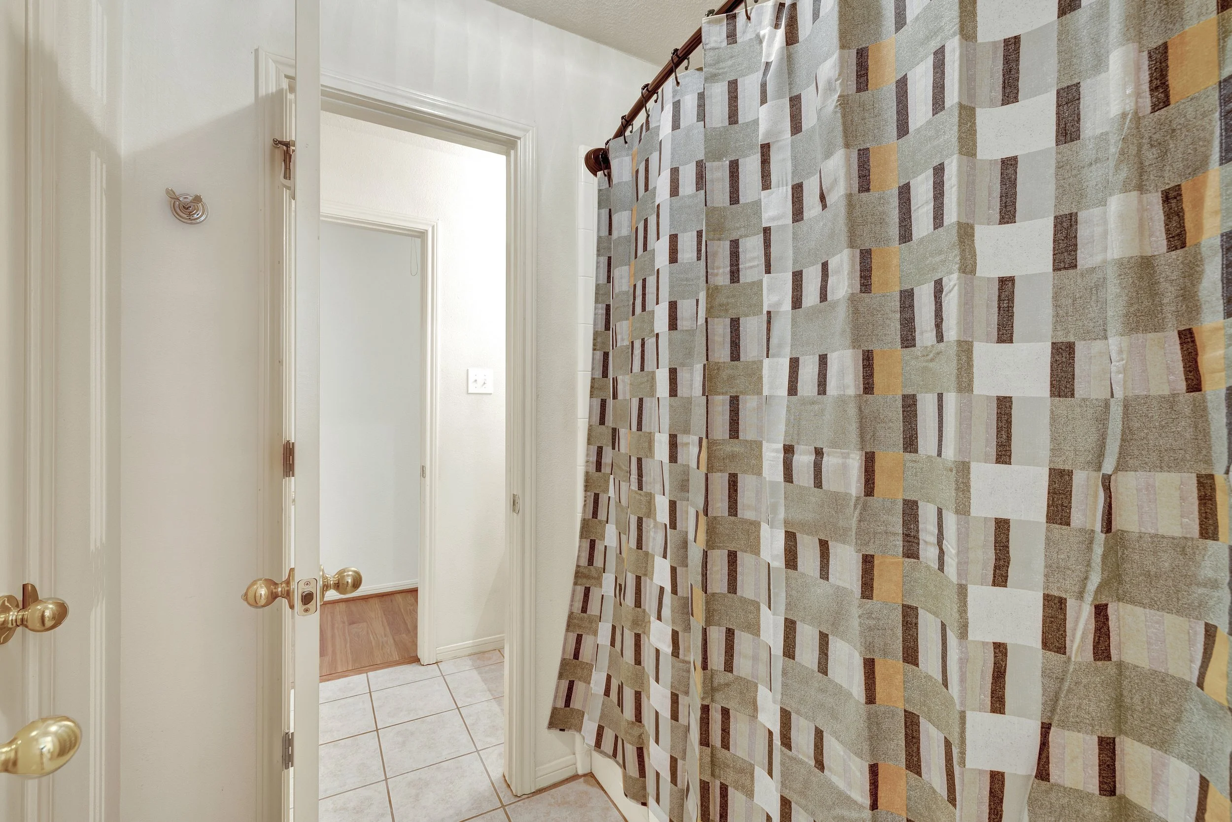 View of a bathroom with a patterned shower curtain, white walls, a door slightly open revealing a hallway with wood flooring, and tiled flooring in the bathroom.