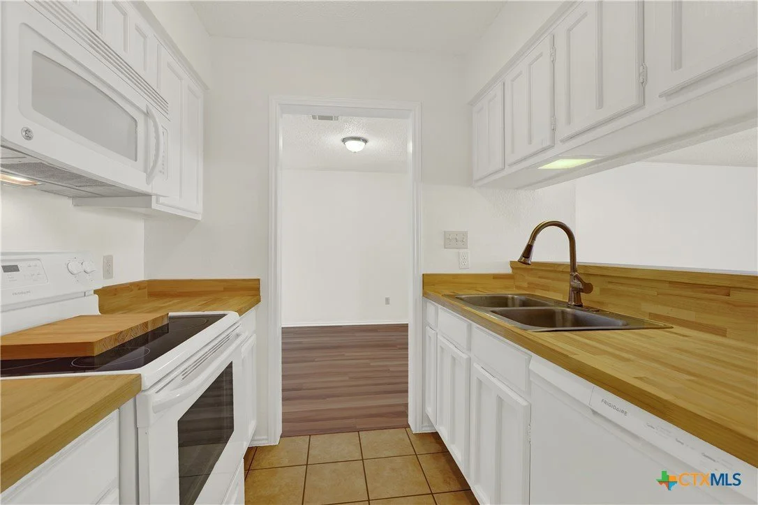 Kitchen with white cabinets, wooden countertops, a double sink, and a white stove. Tiled flooring in front, with a view into an adjacent room with wood flooring.