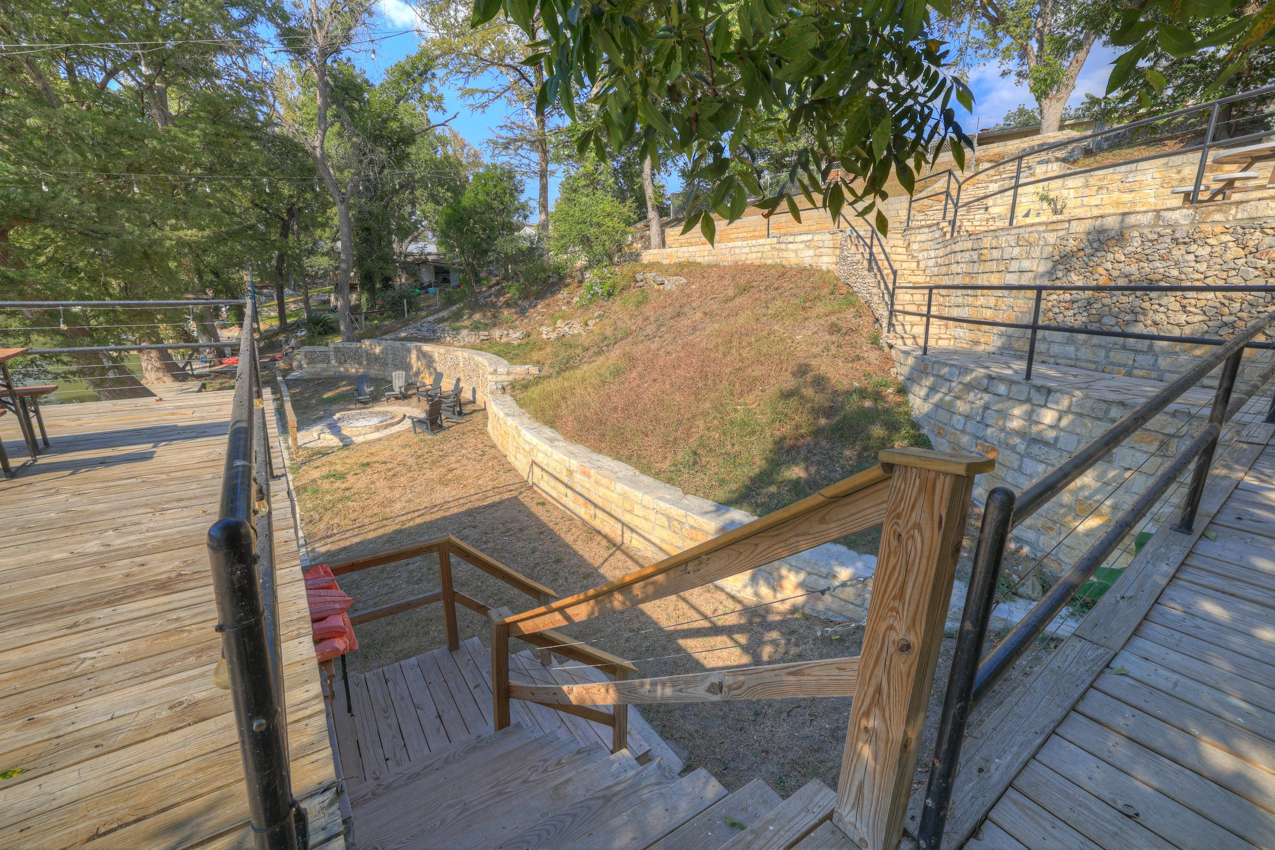 View of a backyard with a wooden deck, stairs, a fire pit area surrounded by chairs, stone retaining walls, grassy slopes, and trees under a partly cloudy sky.
