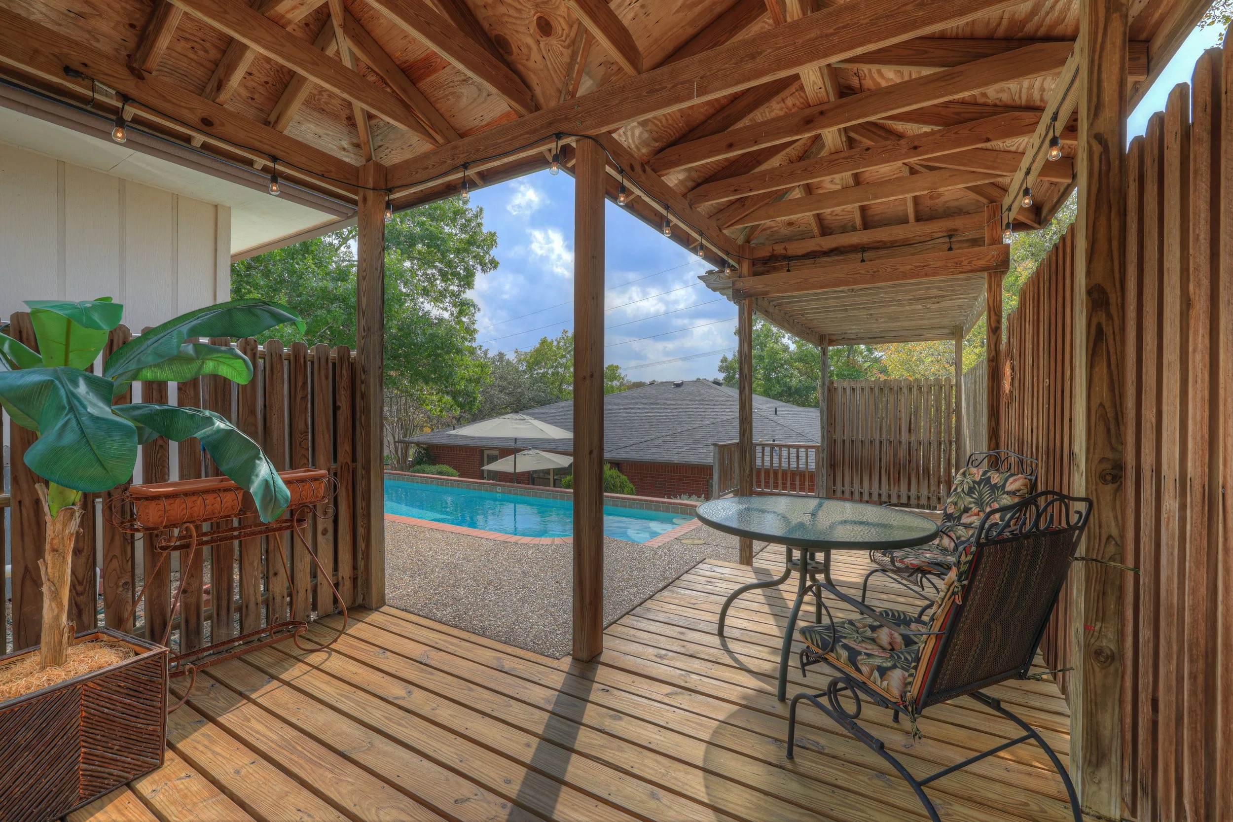 Covered outdoor patio with wooden decking and a seating area with a glass table and floral cushioned chairs, overlooking a backyard with a swimming pool, trees, and neighboring houses.