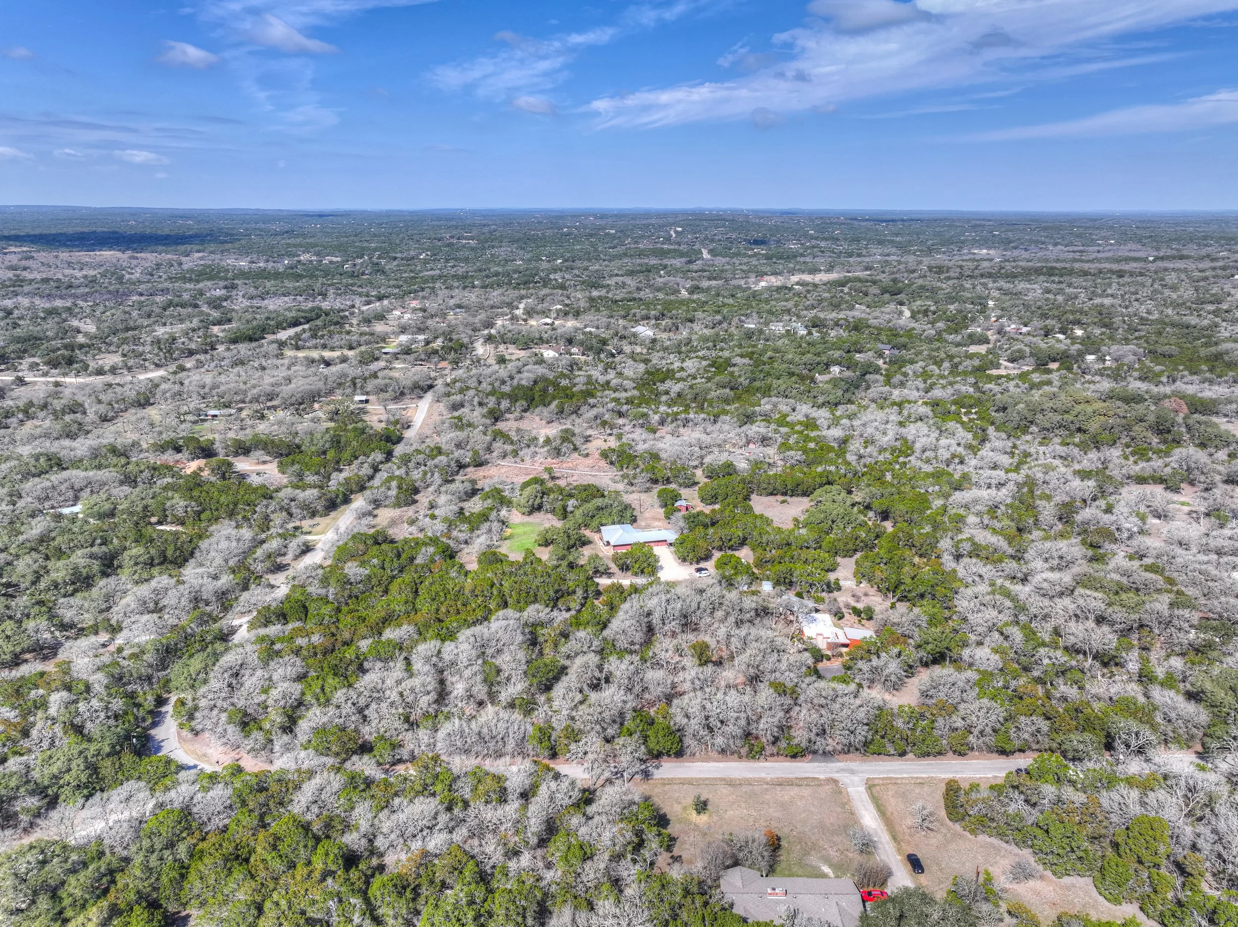 Aerial view of a rural landscape with trees, houses, and roads under a partly cloudy blue sky.