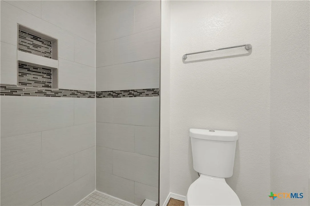 Bathroom with a walk-in shower featuring beige tiles, a strip of small mosaic tiles, and a recessed shelf, next to a white toilet and a towel rack on the wall.