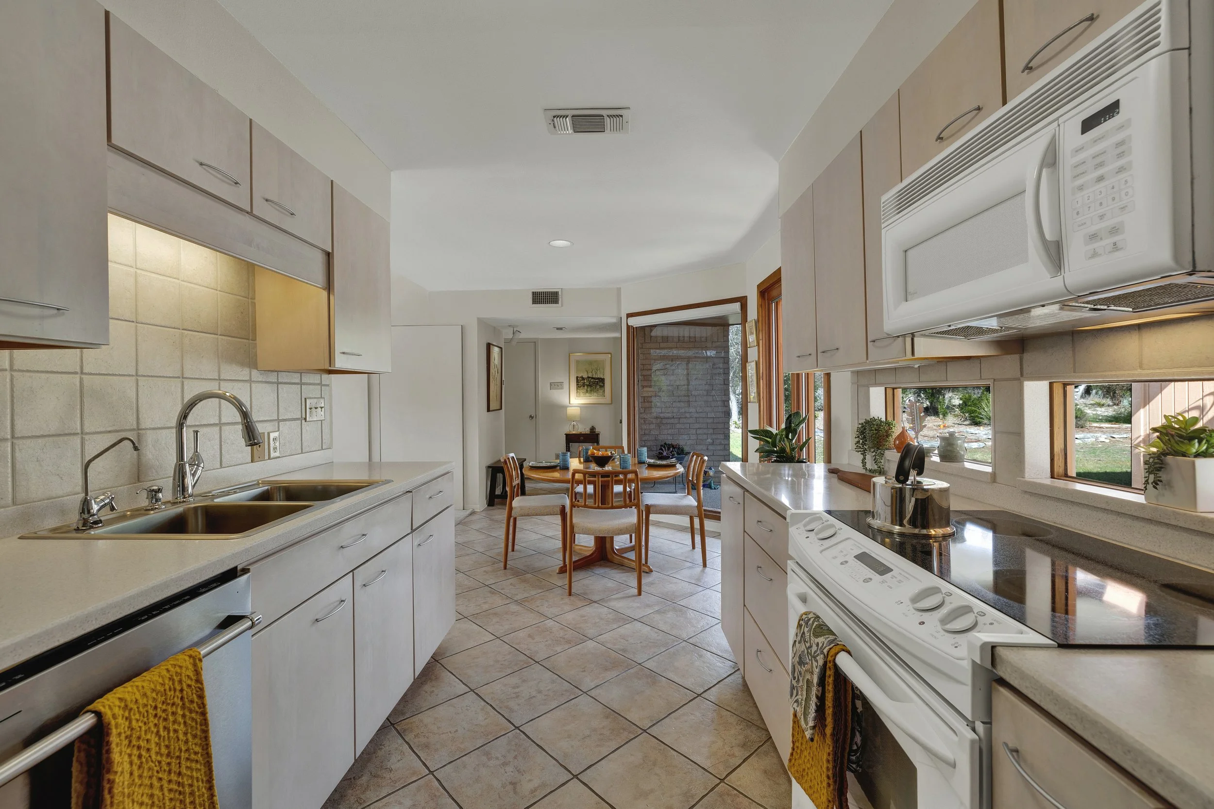 Kitchen with white cabinets, beige tile flooring, window views of a backyard, and a dining area with chairs.