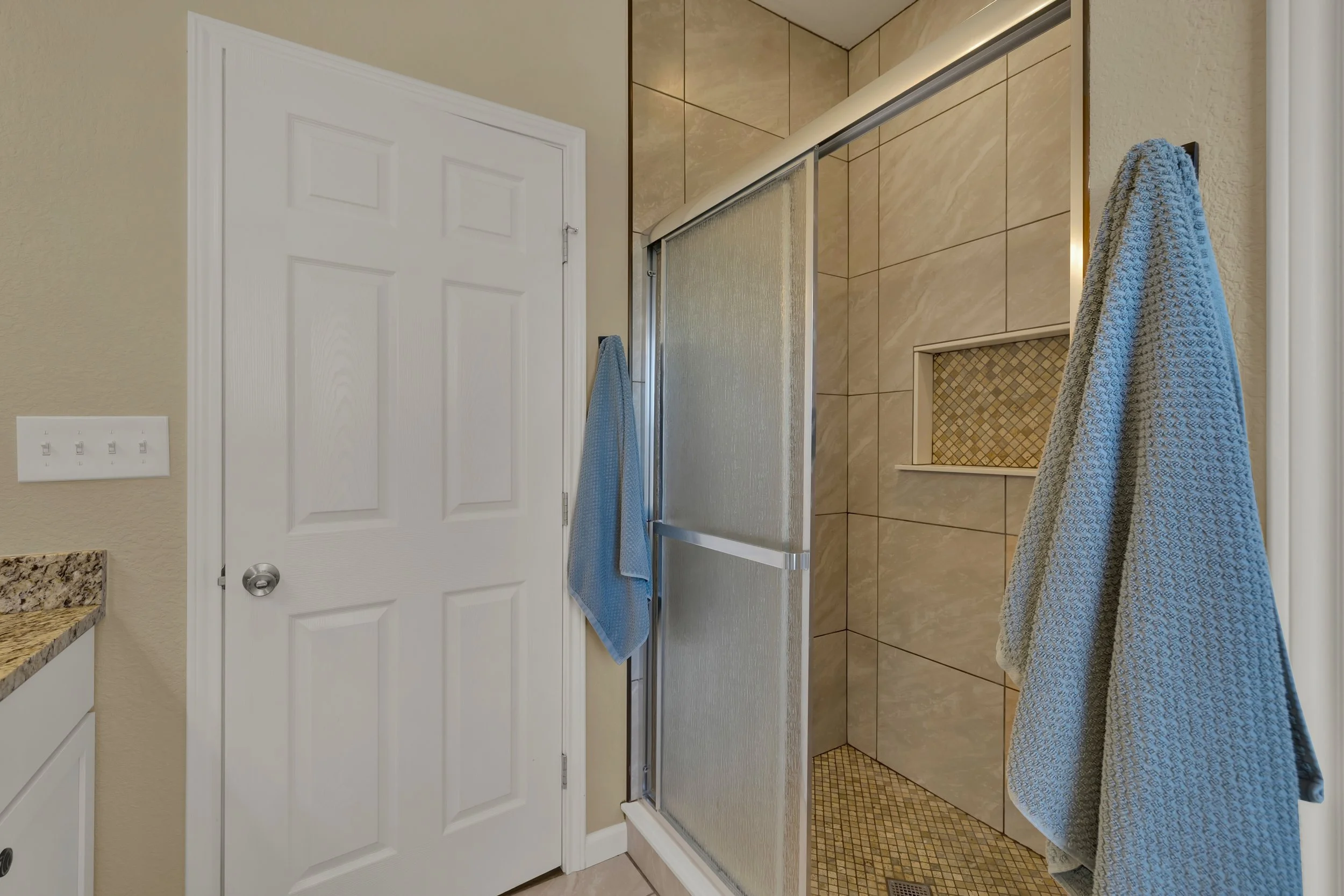 Bathroom shower with beige tile walls, glass door, and built-in niche, with two blue towels hanging on the wall.