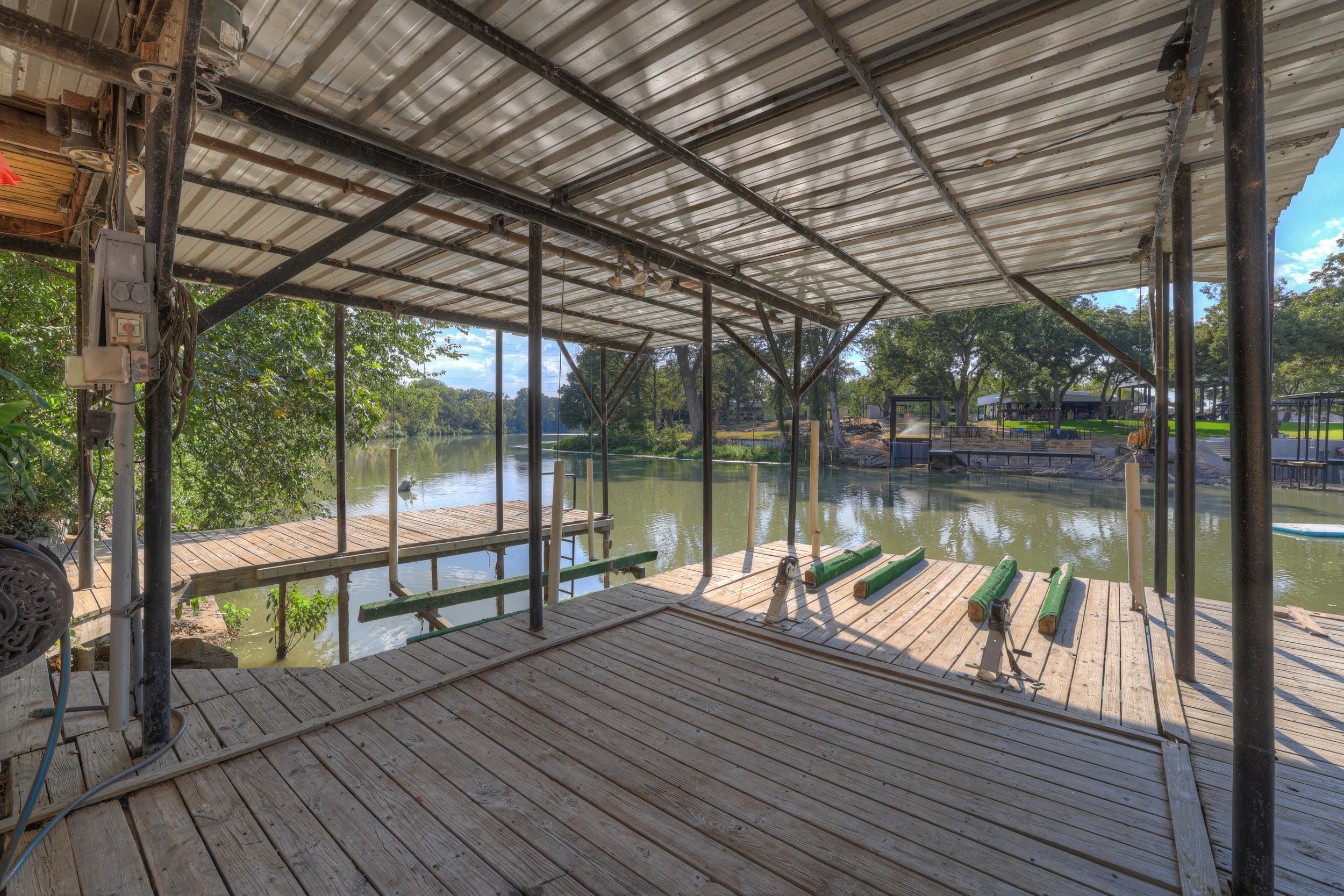 View of a wooden dock with an overhead metal roof by a river, with trees and a park in the background.
