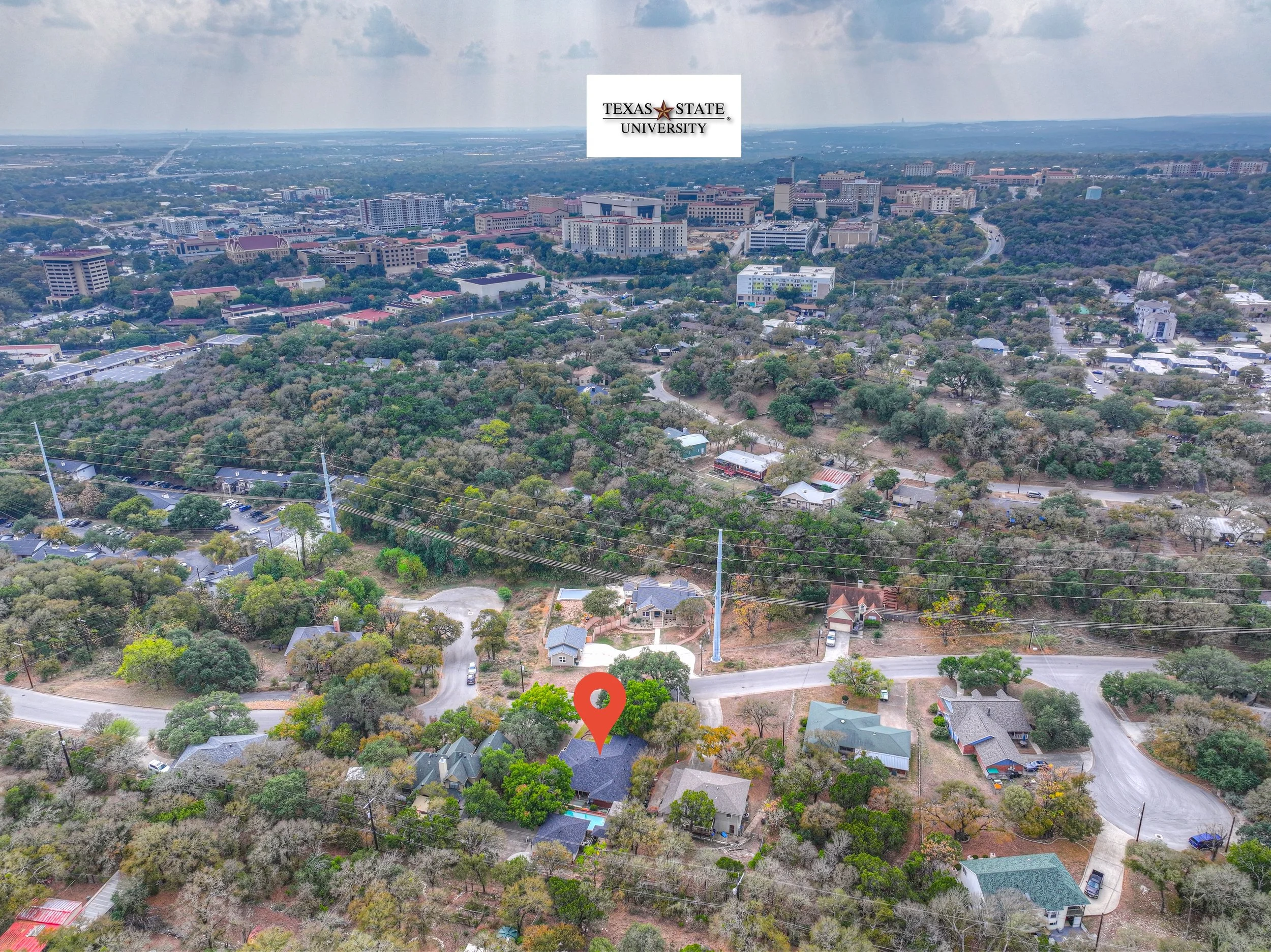 Aerial view of a suburban neighborhood with trees, houses, and roads, with Texas State University campus in the distance. The campus is at the top center of the image, with multiple buildings and a large parking lot, under a partly cloudy sky.