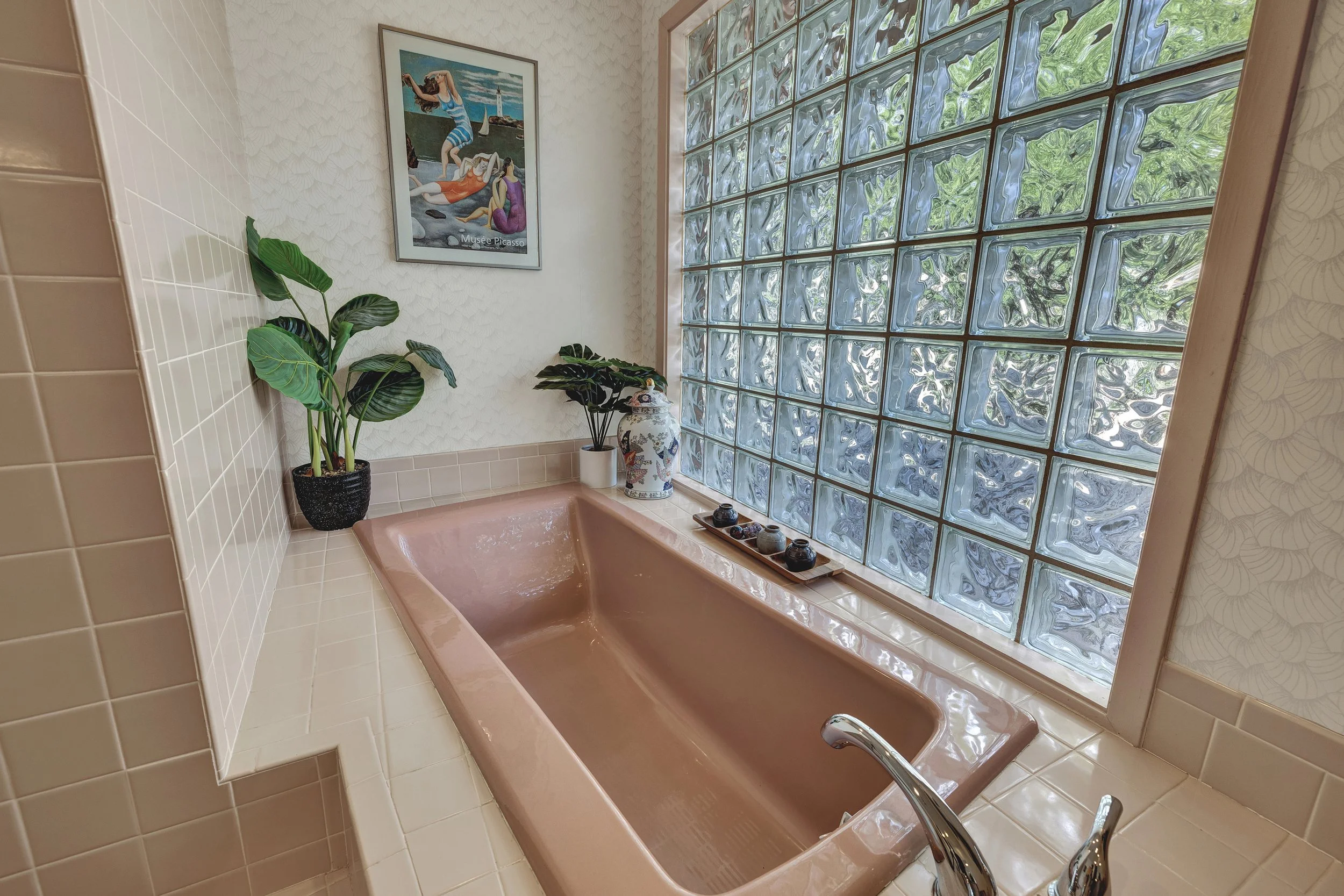 A pink bathtub in a bathroom with decorative plants, a vase, artwork on the wall, and a large glass block window letting in natural light.