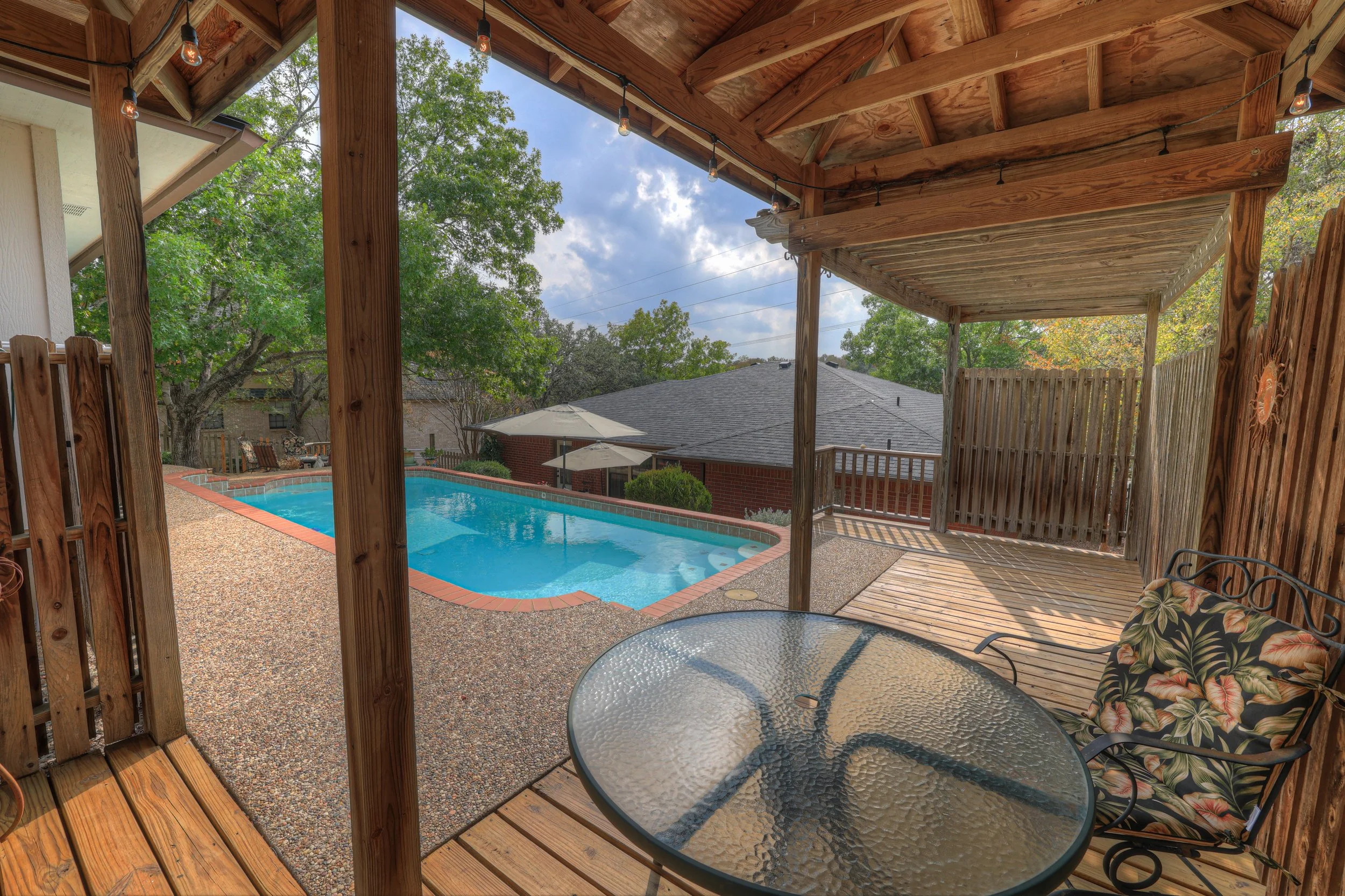 Covered wooden patio with glass-topped table and floral chair cushion overlooking a backyard pool with umbrella-shaded seating area and trees.