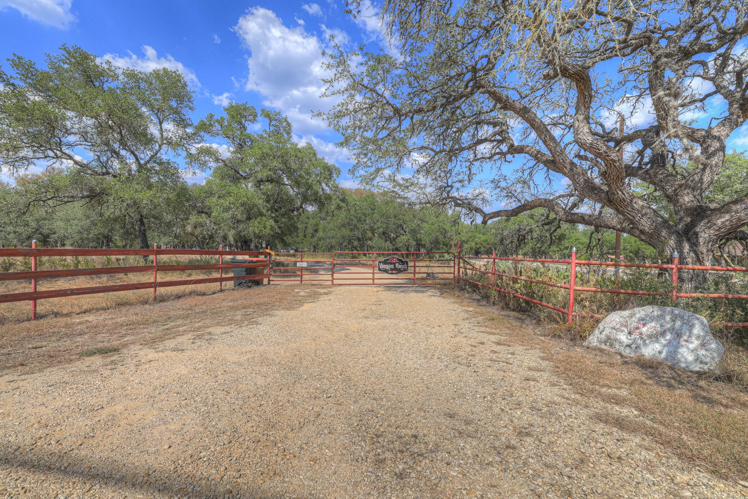 Entrance to Vazquez Ranch with a gravel path, red metal fence, large tree on the right, and a sign on the gate, under a partly cloudy blue sky.