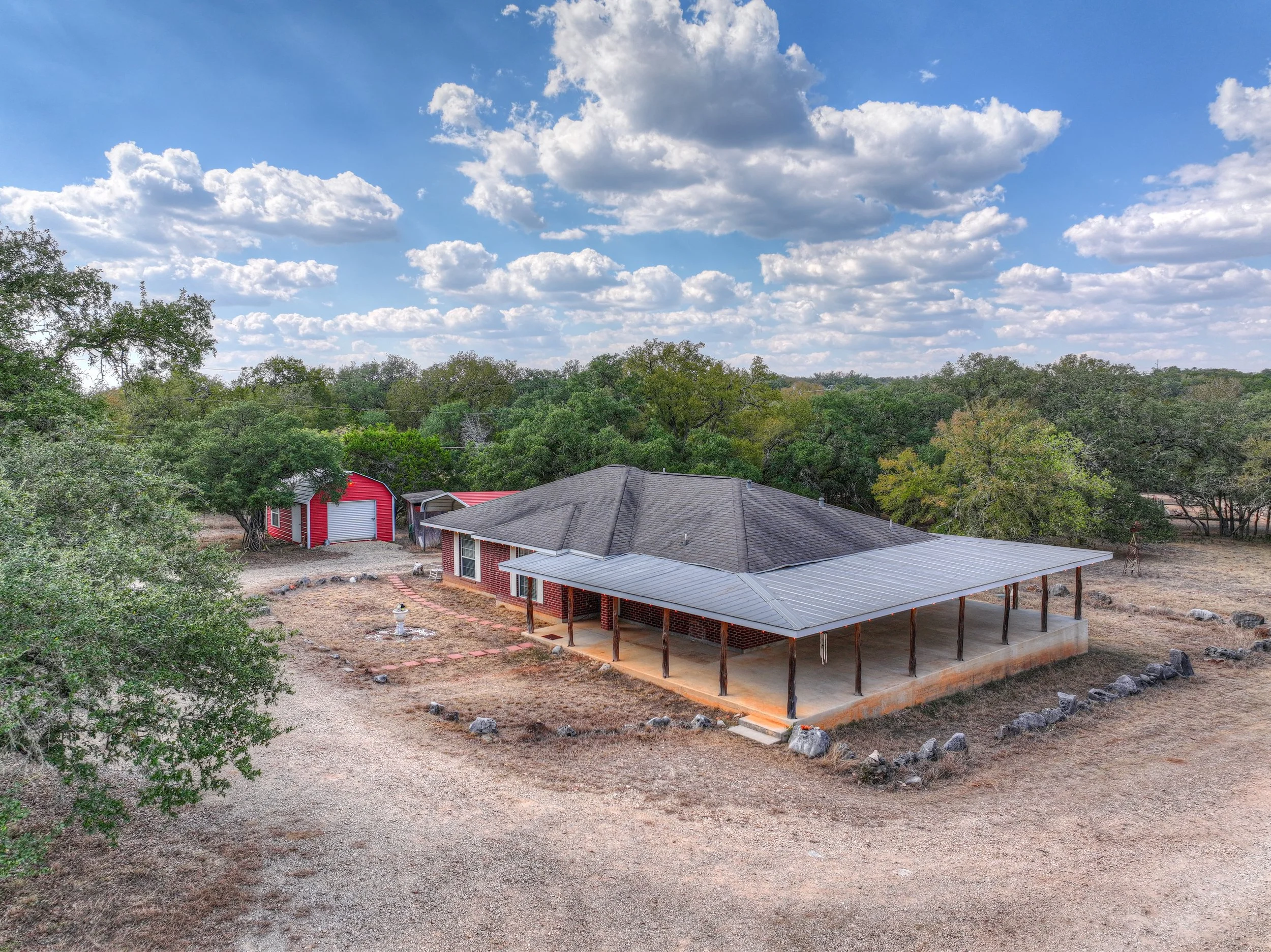 A house with a covered porch and gray roof, a red barn with a white door, surrounded by trees, under a blue sky with scattered white clouds.