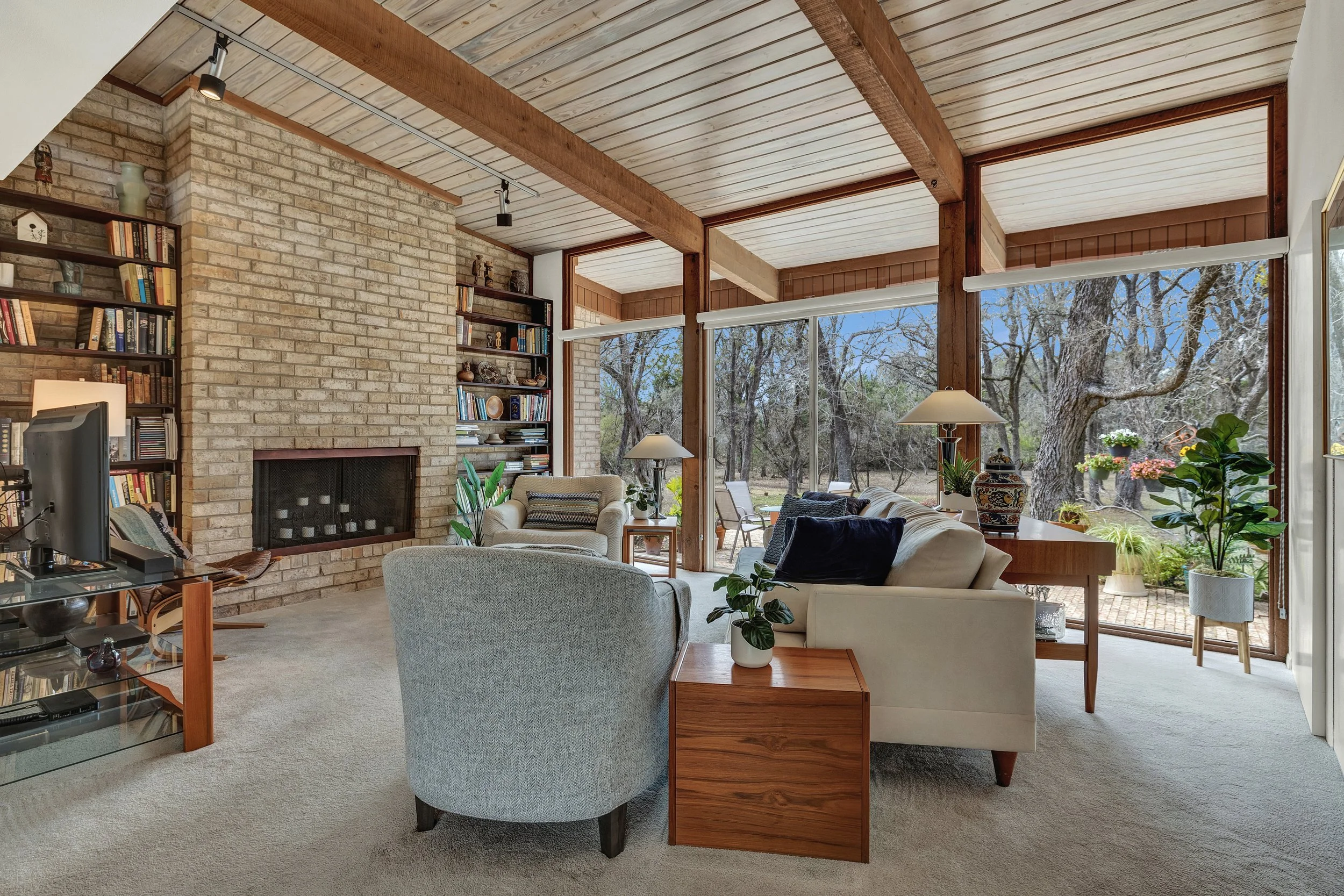 Living room with brick fireplace, large windows with view of trees, beige carpet, wooden beams and ceiling, and various potted plants and furniture.