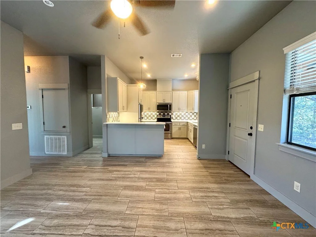 Empty living area with wooden floors, gray walls, a ceiling fan, and a view into a kitchen with white cabinets and a patterned backsplash.