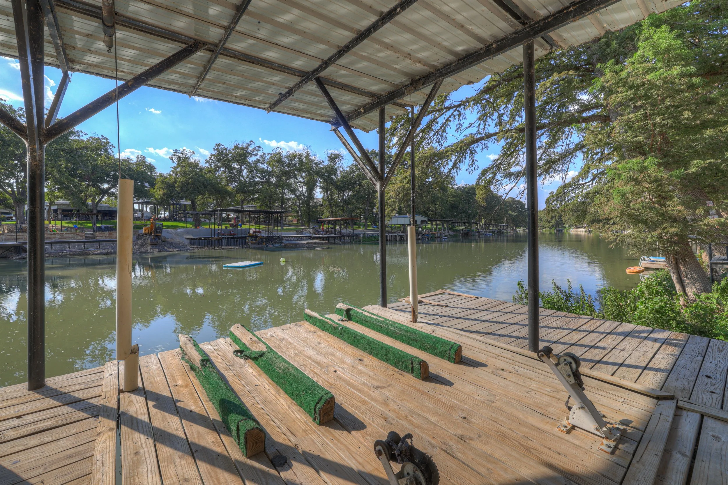 Empty boat dock with wooden planks, metal framework, and green artificial turf, by a calm river with trees and boats in the background under a blue sky with clouds.