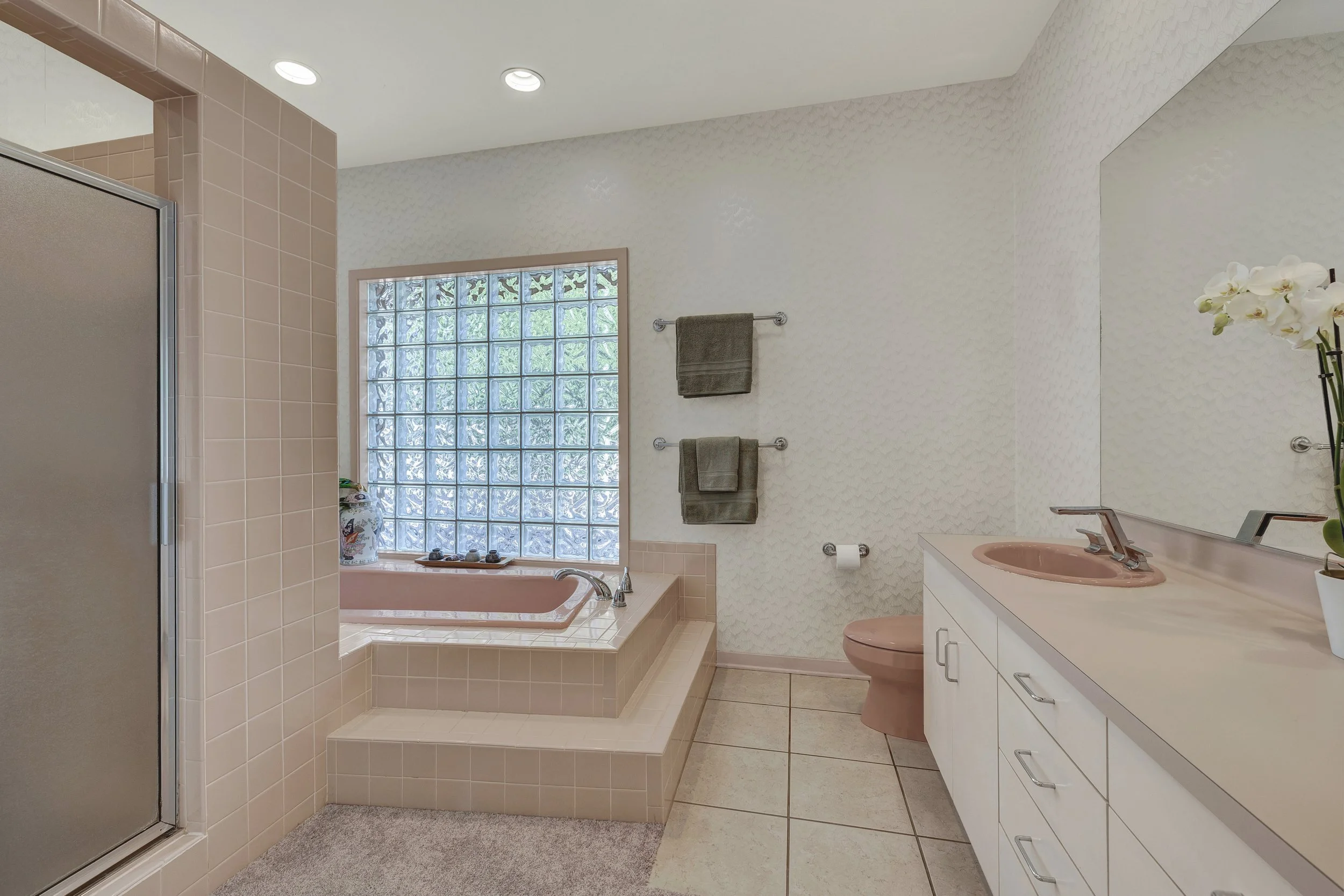 A bathroom with a pink bathtub and sink, a large mirror, a glass brick window, green towels on a towel rack, a toilet, and a vanity with storage drawers.