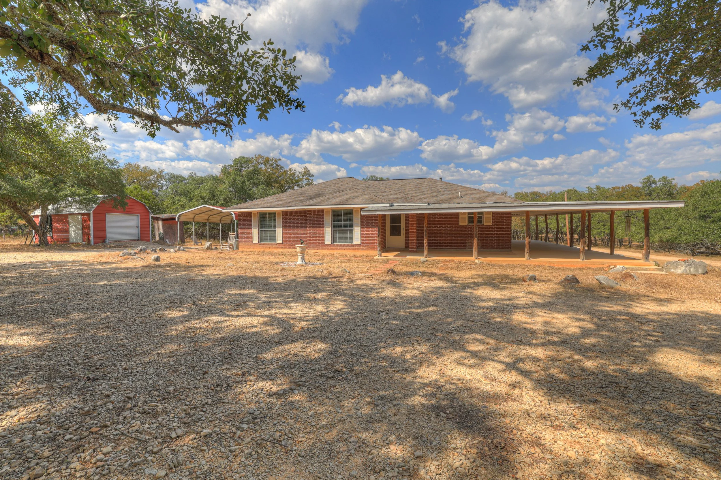 A brick house with a covered backyard porch and a separate red shed, surrounded by trees and a gravel yard under a partly cloudy sky.