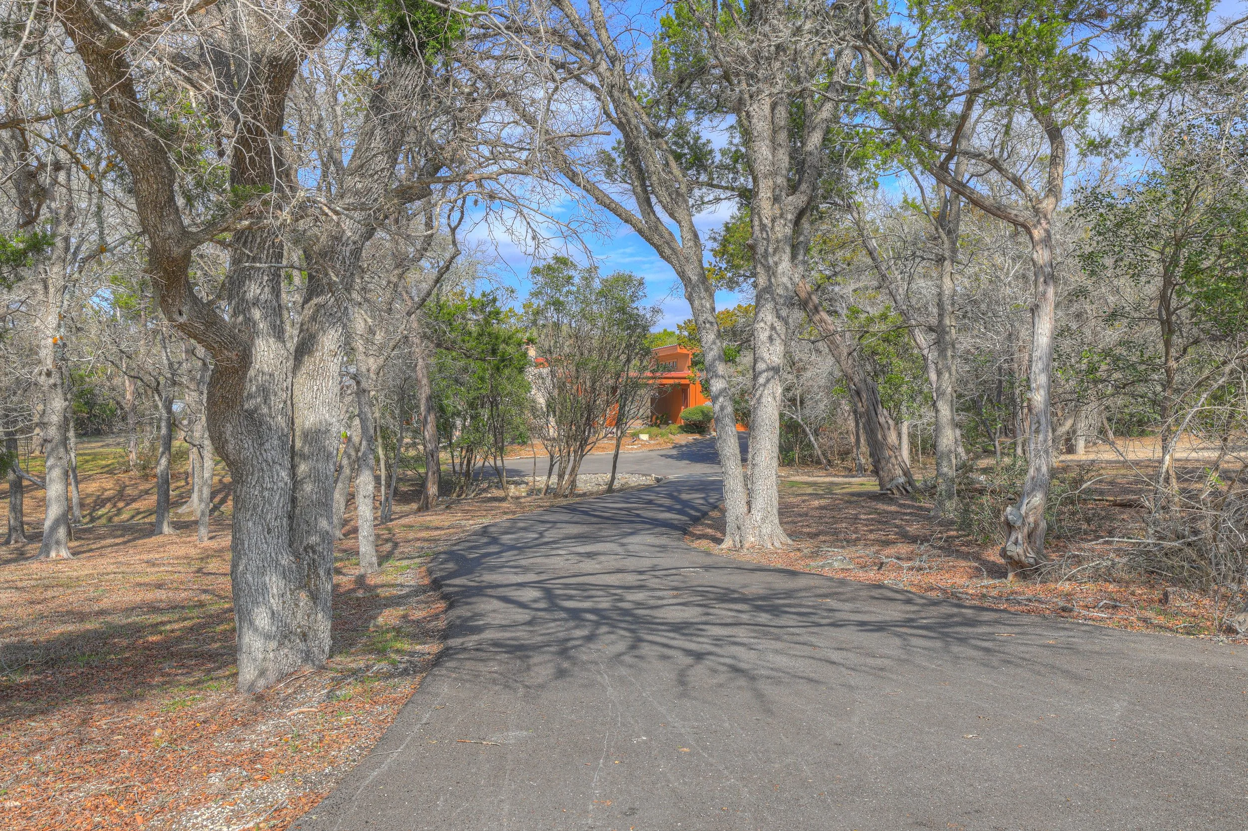 A paved driveway winding through a wooded area with leafless trees and some green foliage. A house with an orange exterior is visible at the end of the driveway.
