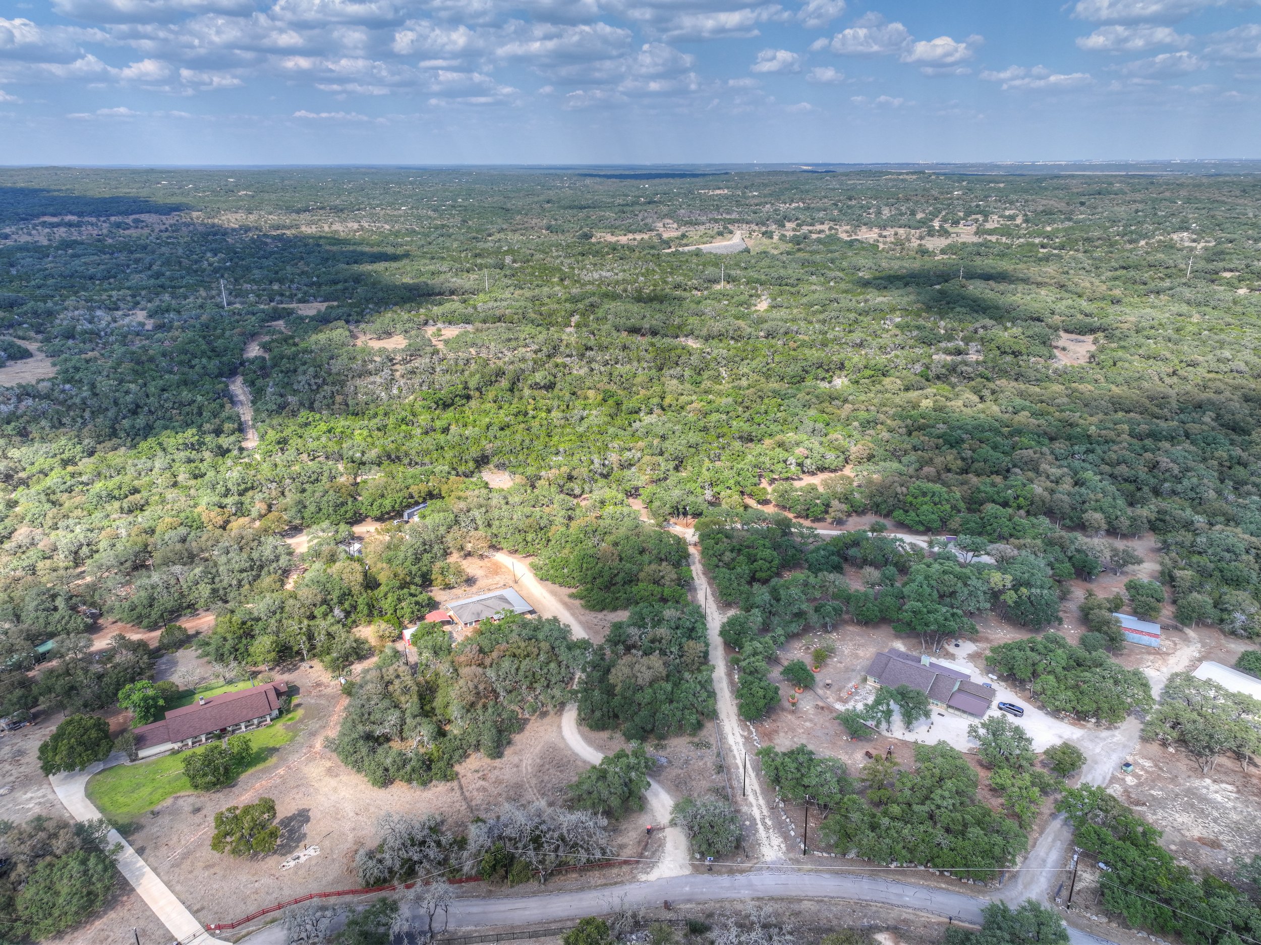Aerial view of a rural area with dense green trees, scattered houses, and unpaved roads. The landscape extends to the horizon under a partly cloudy sky.