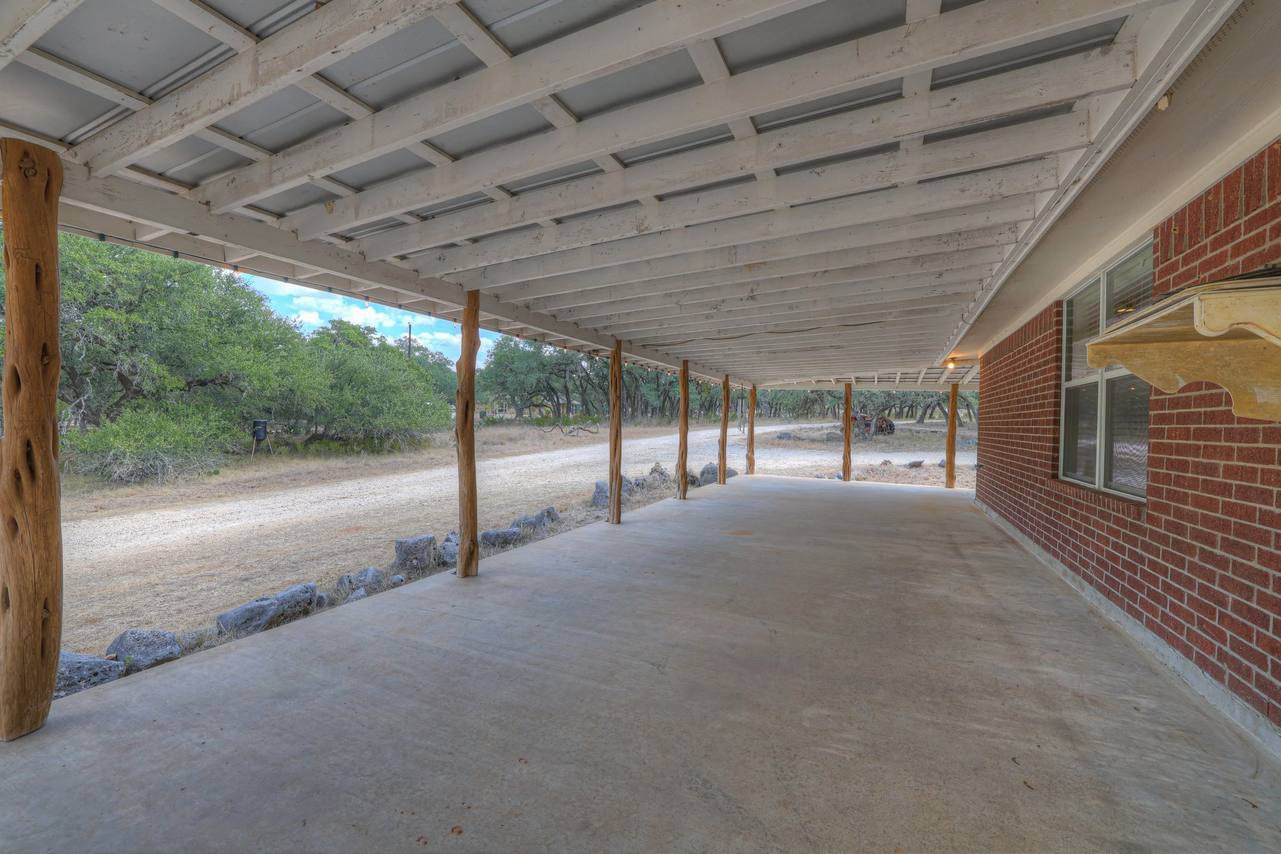 Outdoor patio with a concrete floor, supported by wooden posts, and an overhanging roof. There is a brick house wall with a window on the right, and a landscape with trees and dirt road in the background.