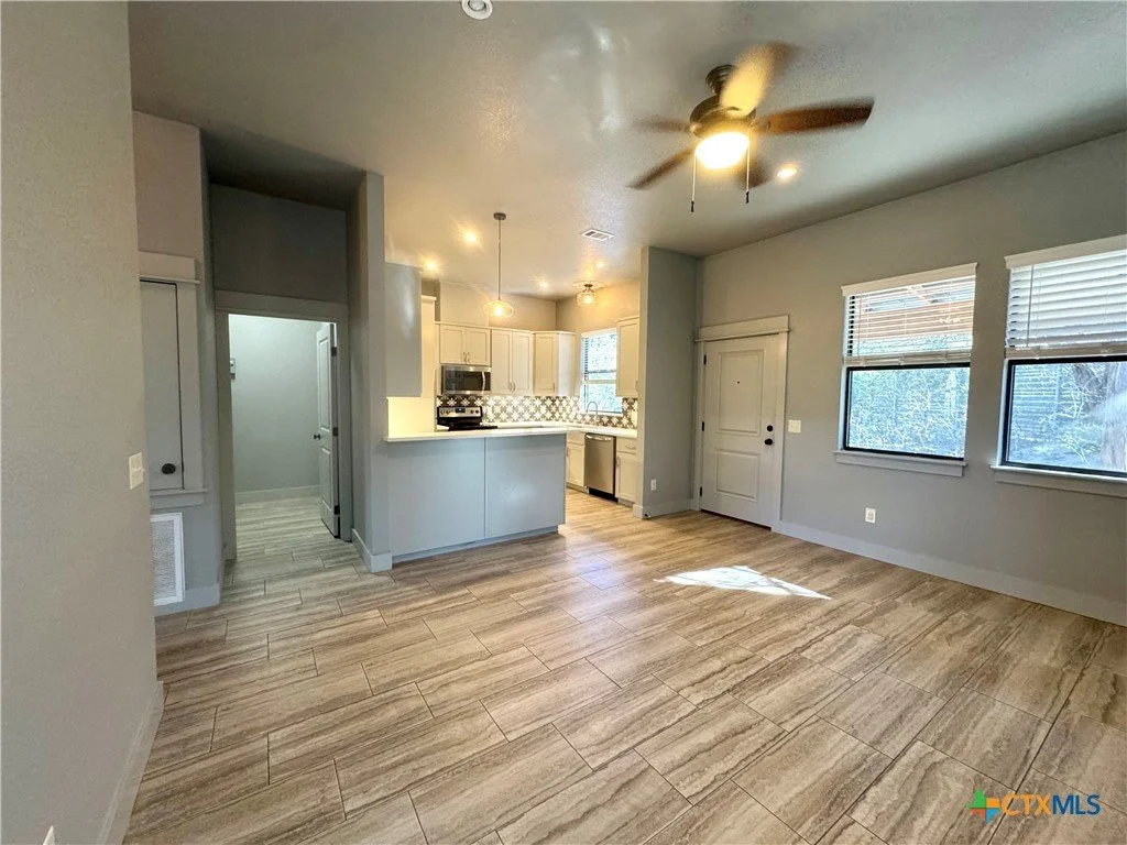 Empty living room with a ceiling fan, wooden floors, and three windows. An open kitchen with white cabinets, a counter, and appliances is visible in the background.