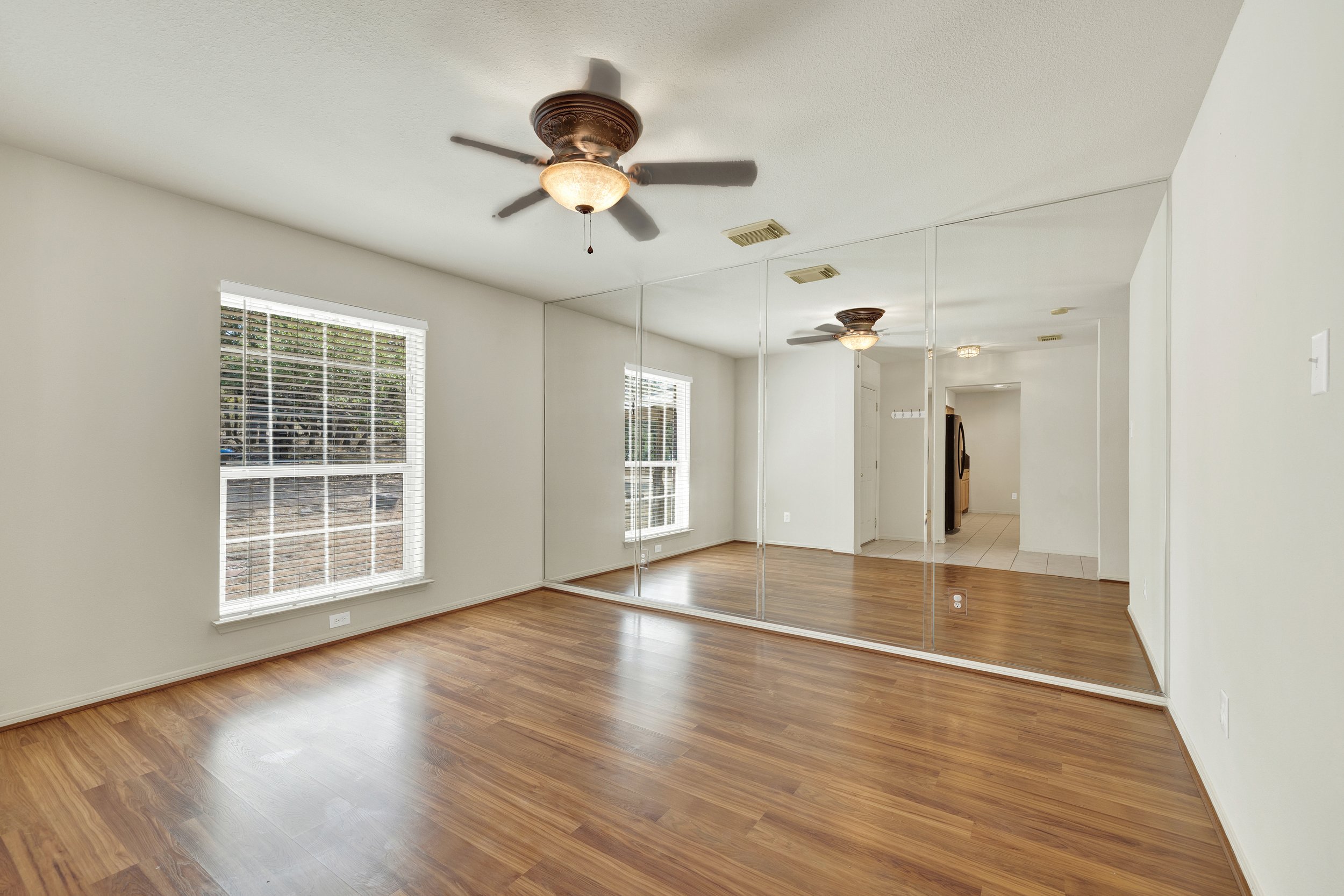 Empty living room with hardwood floors, large windows with blinds, mirrored closet doors, ceiling fans, and a doorway leading to a kitchen area.
