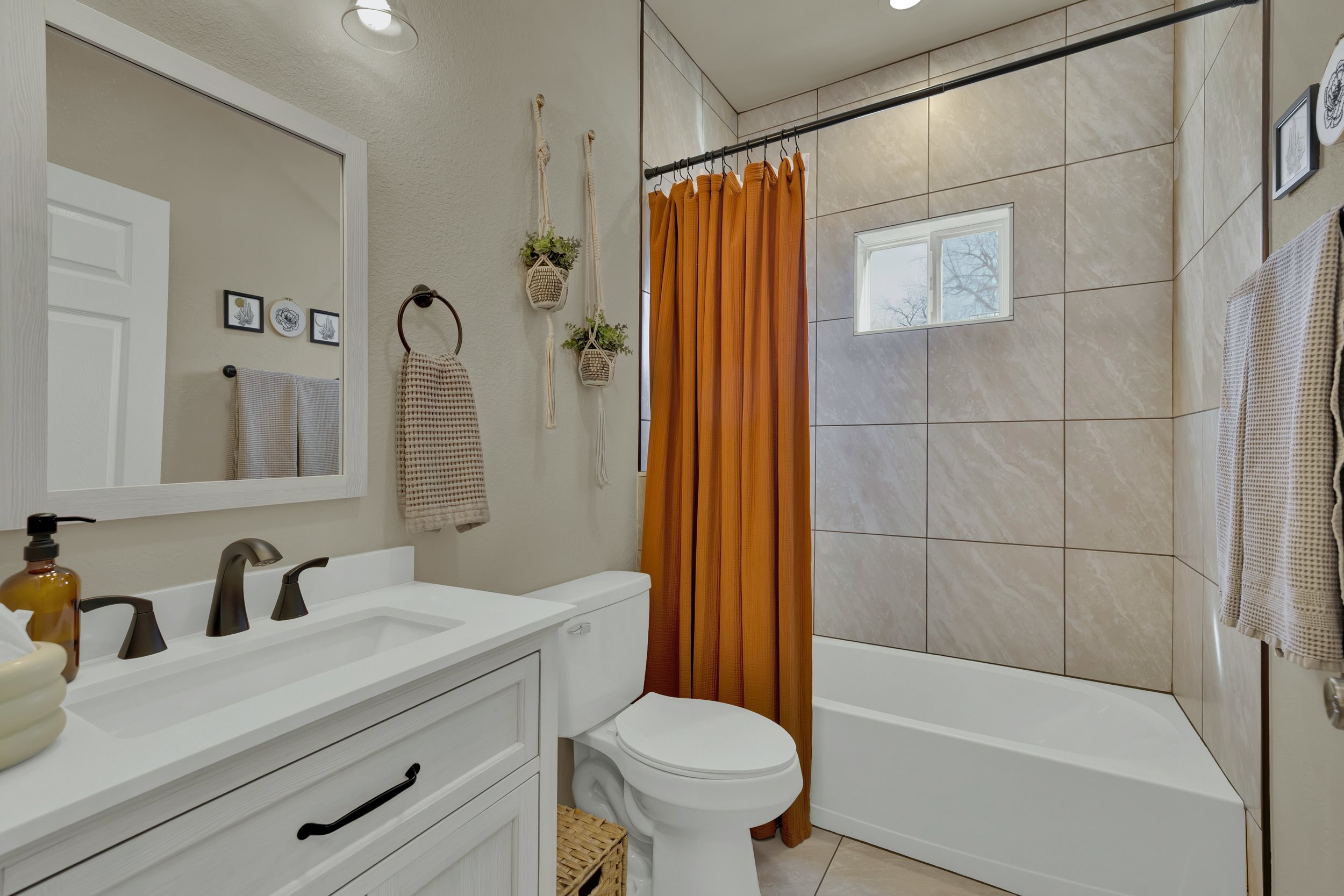 Bathroom with white vanity, brown faucet, mirror, toilet, beige tiles, small window, orange shower curtain, and decorative hanging plants.