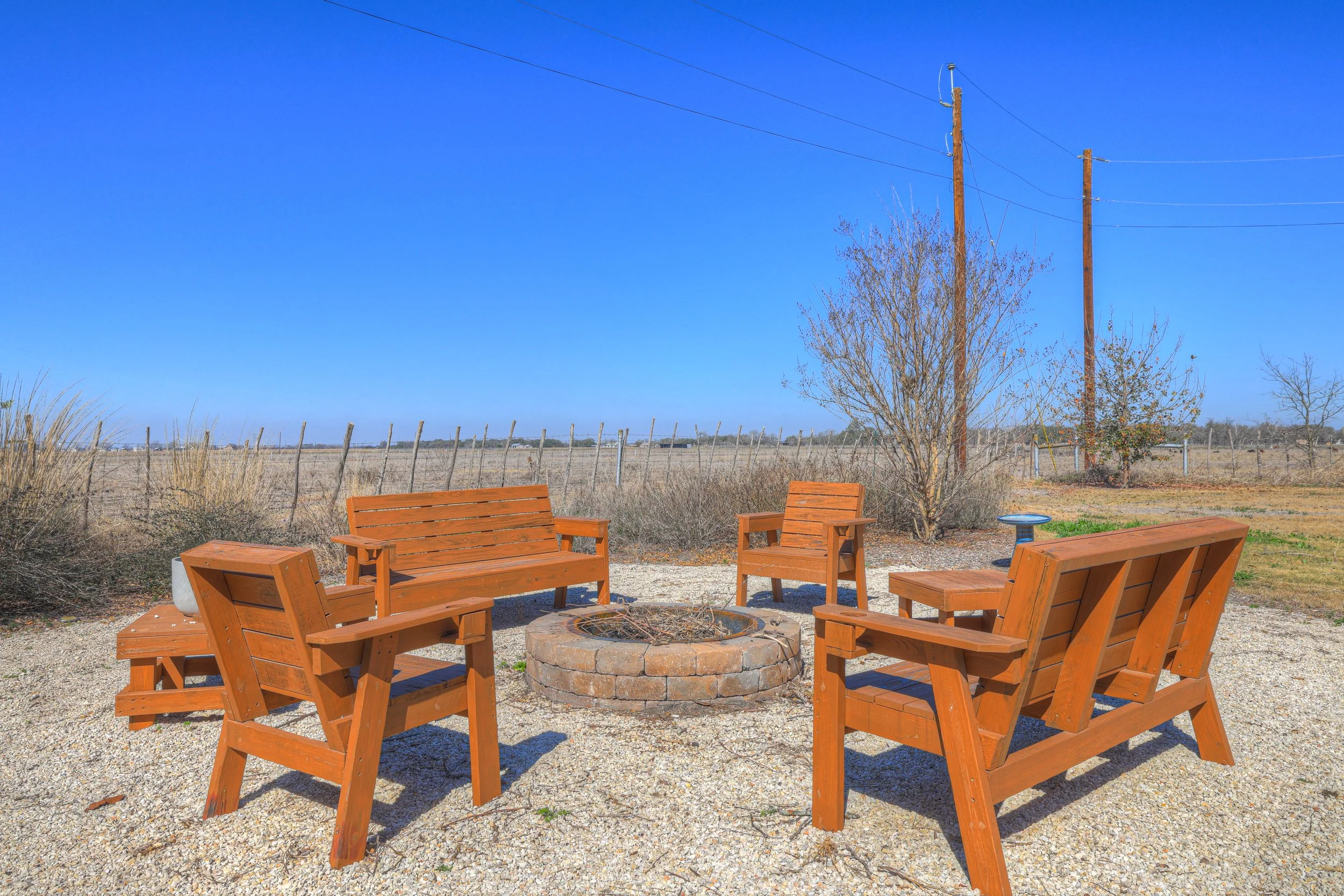 Four wooden outdoor benches arranged around a firepit in a rural, open field under a clear blue sky.