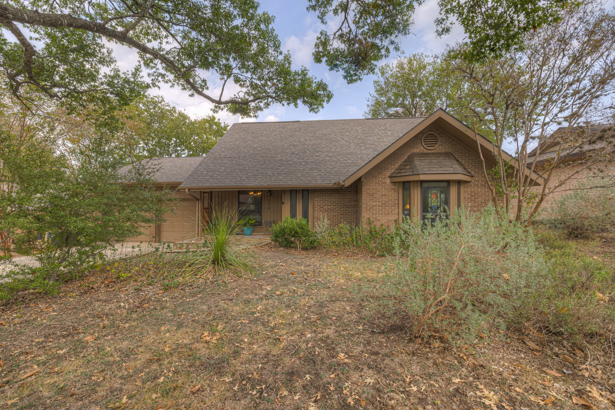 A single-story brick house with a sloped roof, front door, and a bay window, surrounded by trees and bushes, in a dry, leaf-strewn yard.